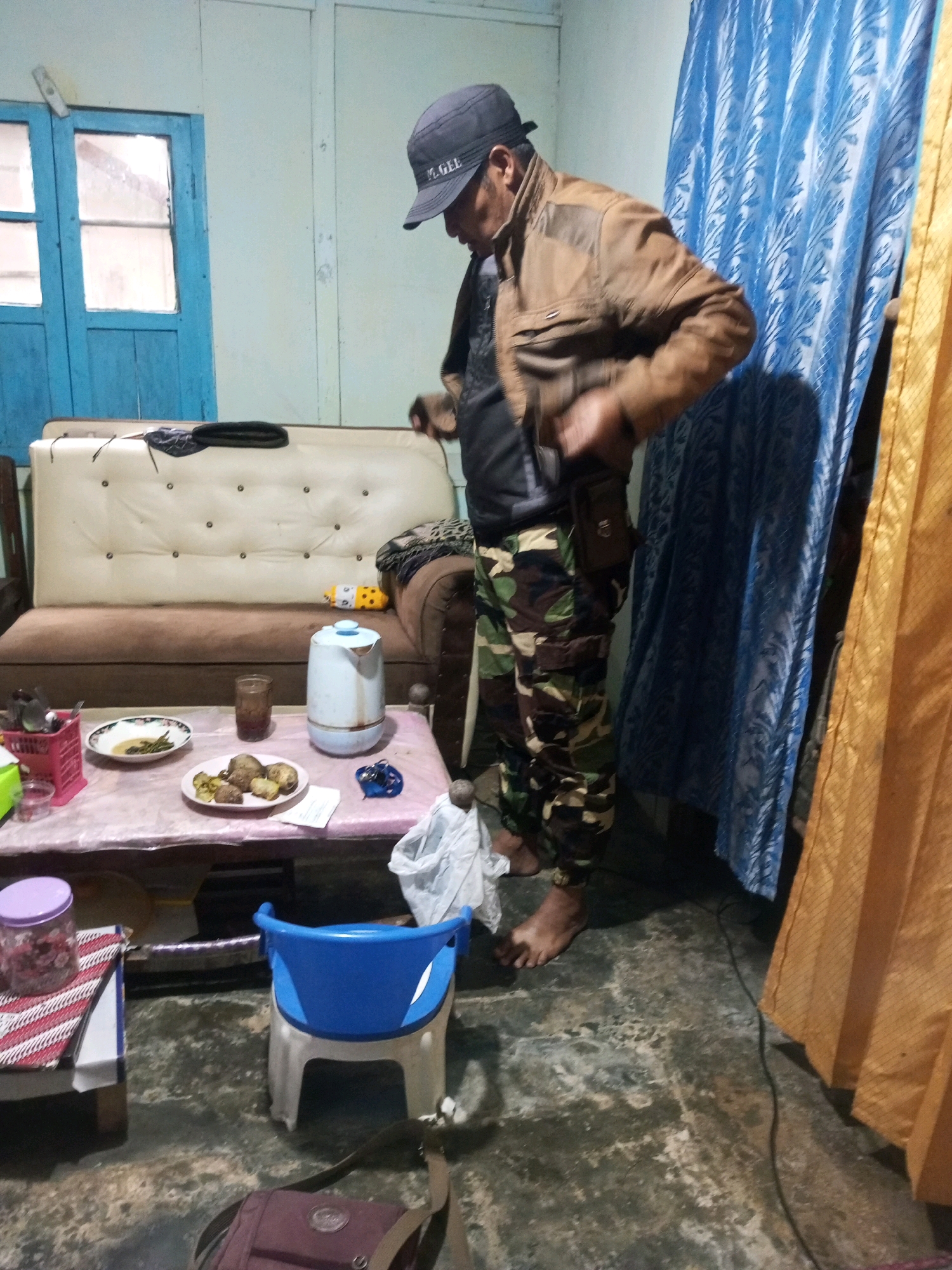 A person wearing a brown jacket and camouflage pants stands in a room, adjusting their clothing. To the left is a sofa with a cream-colored upholstered back and brown lower cushions. In front of the sofa is a table covered with a pink plastic sheet, holding plates of food, a glass, and a white electric kettle. A small blue chair sits to the right of the table. The room has light blue walls and a blue and yellow patterned curtain to the right. The floor appears to be concrete. The scene is consistent with a domestic setting in Andongsili, Indonesia.