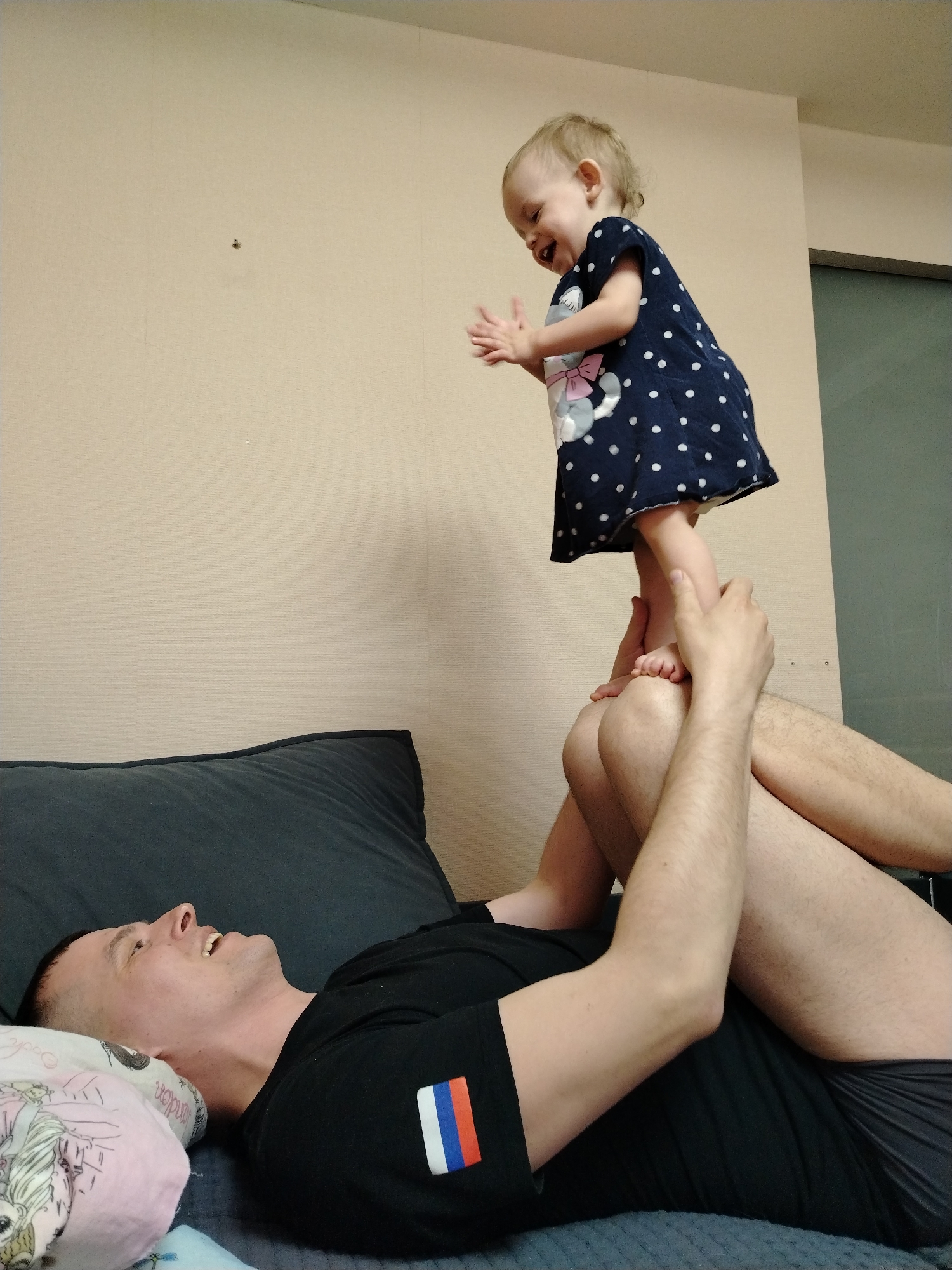 A man and a young child are indoors. The man lies on his back on a bed with a gray headboard and a pillow with a cartoon design. He is wearing a black t-shirt with the Russian flag on the left sleeve and dark shorts. The child stands on the man's feet as he holds her legs. She wears a navy blue dress with white polka dots and a cat design. The child is smiling and clapping her hands. The wall is beige with a small spot. The location is presumably a residence in the city of Chaikovsky, Russia.