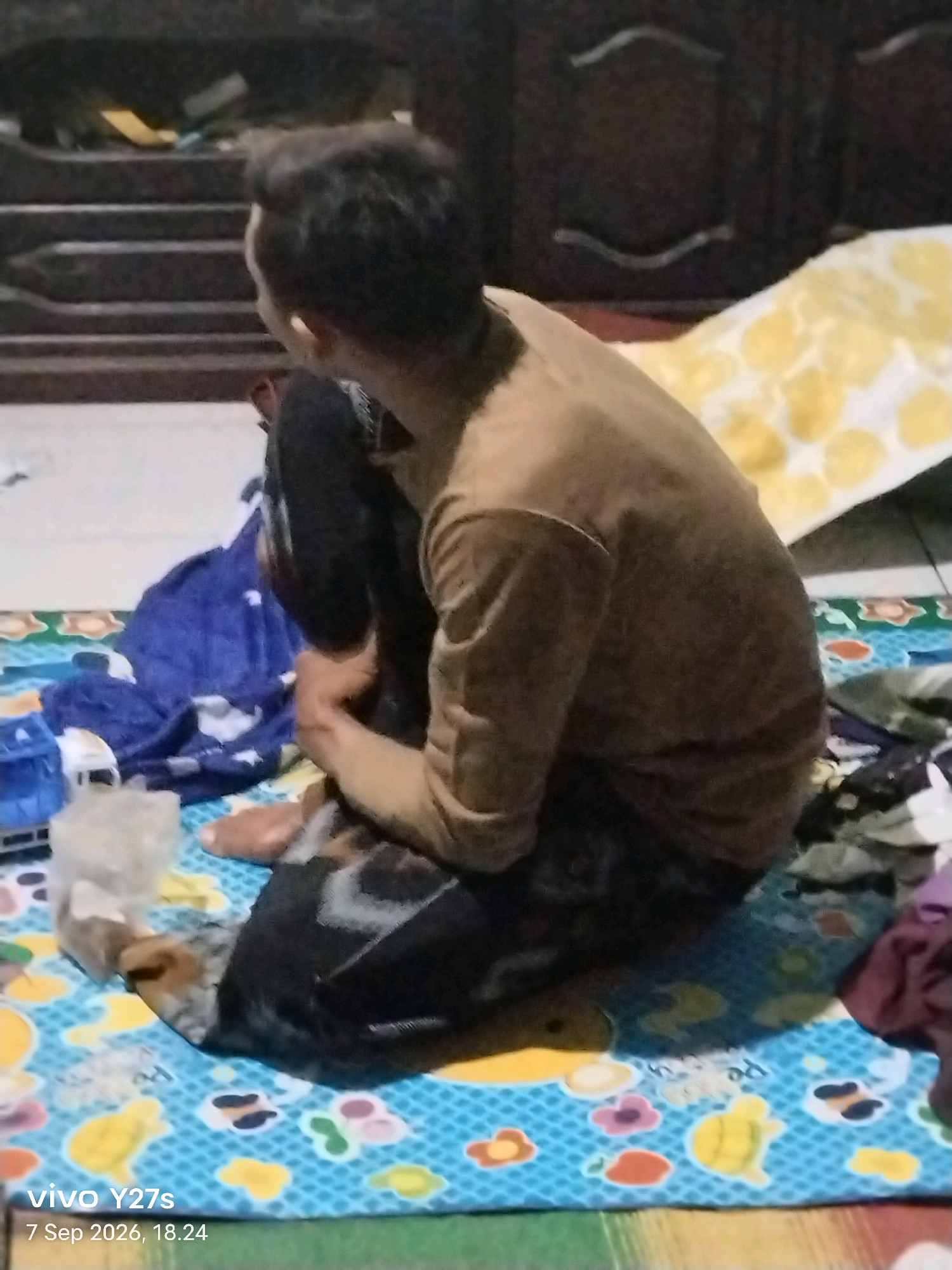 A young man is sitting on a blue mat with a yellow and white patterned blanket in the background. He is wearing a brown shirt and black pants, and his legs are crossed. A blue blanket is spread out on the mat to his left. A small blue toy is visible on the mat as well. The image appears to have been taken indoors, possibly in a residential setting. The lighting is dim, suggesting it might be evening or a poorly lit room. A watermark in the bottom left corner indicates the image was taken on "7 Sep 2026, 18.24" with a "vivo Y27s" device. There is no specific indication of the location being in the city of Tamansari, Indonesia.