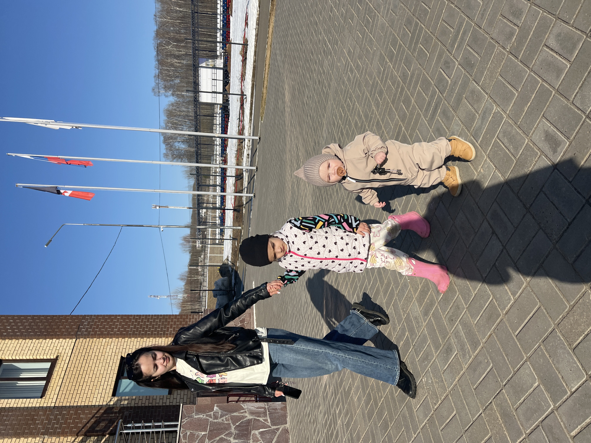 The image shows a woman and two children on a paved surface in Noviy, Russia. The woman, wearing a black leather jacket, blue jeans, and black shoes, holds the hand of a child wearing a white patterned jacket, black hat, and pink boots. A second child, dressed in a beige outfit and hat, lies on the pavement nearby. In the background, there are flagpoles with flags, trees, and a building with red seating. The paving is arranged in a herringbone pattern. The building in the background is likely related to the ice arena.