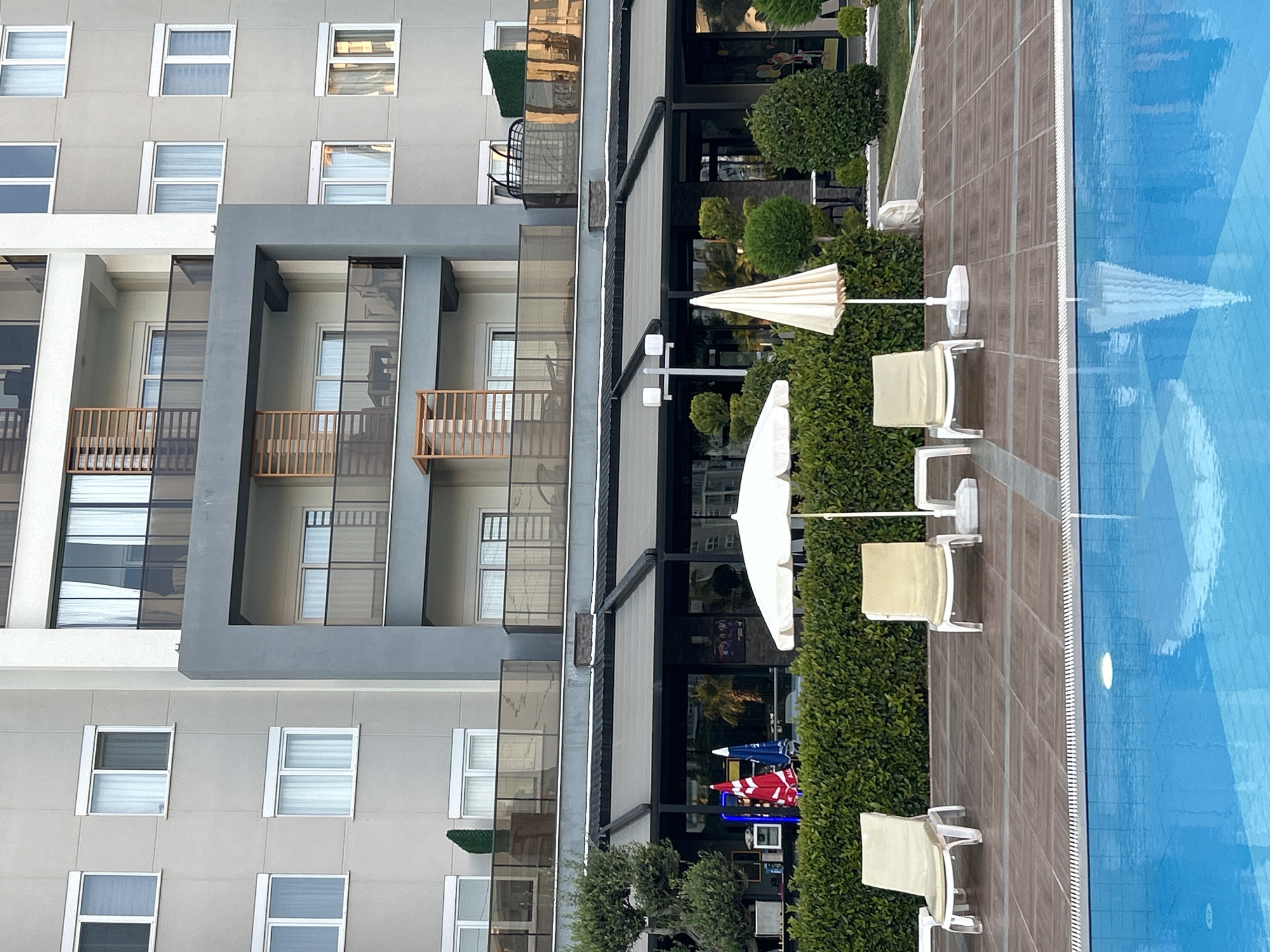 The image captures a scene near a swimming pool in Alanya, Turkey, adjacent to a multi-story building with a modern architectural design. The building features numerous windows and balconies with glass railings. Along the poolside, there are several white sun loungers and closed white umbrellas, positioned on a tiled surface. A line of green bushes separates the lounging area from the building's structure. Part of the pool's blue water is visible on the right side of the image.