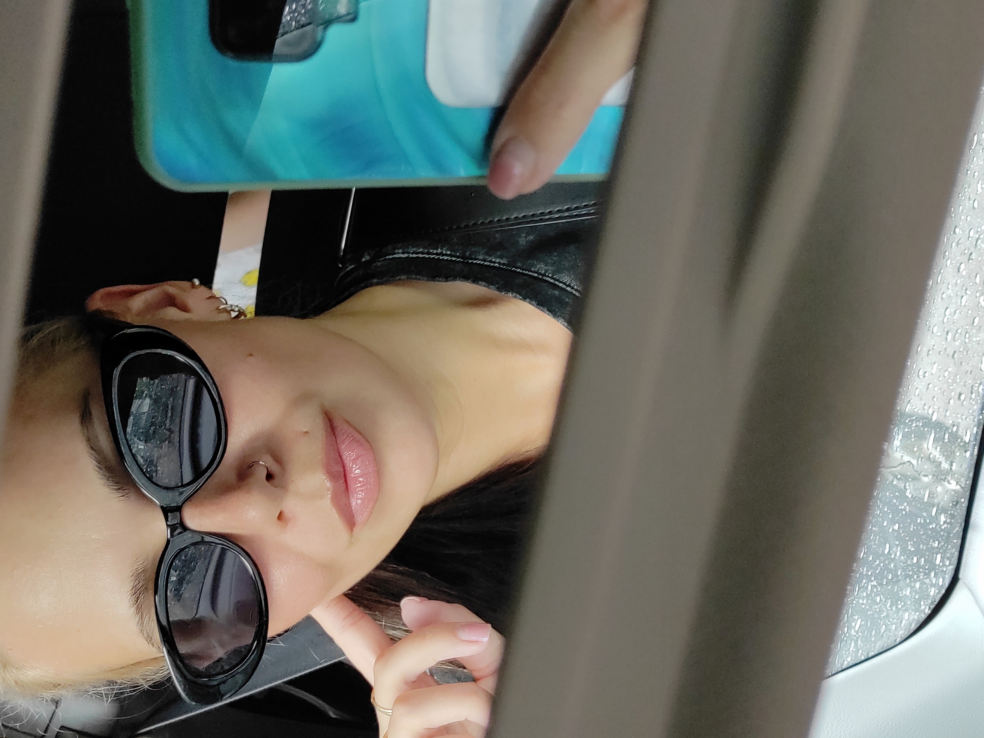 A young woman with blonde hair and fair skin is reflected in a car's rearview mirror. She is wearing black, oval-shaped sunglasses, a nose ring, and gold hoop earrings. Her lips are glossy and pink. She is holding her right hand to her temple, with a gold ring on her ring finger. She is wearing a black, textured jacket. In her right hand, she holds a smartphone with a turquoise and green case. The reflection shows water droplets on the exterior of the car window, indicating it is raining. The background of the reflection is dark, suggesting the interior of the car. The setting appears to be within a city environment, potentially Chaikovsky, Russia, given the context.