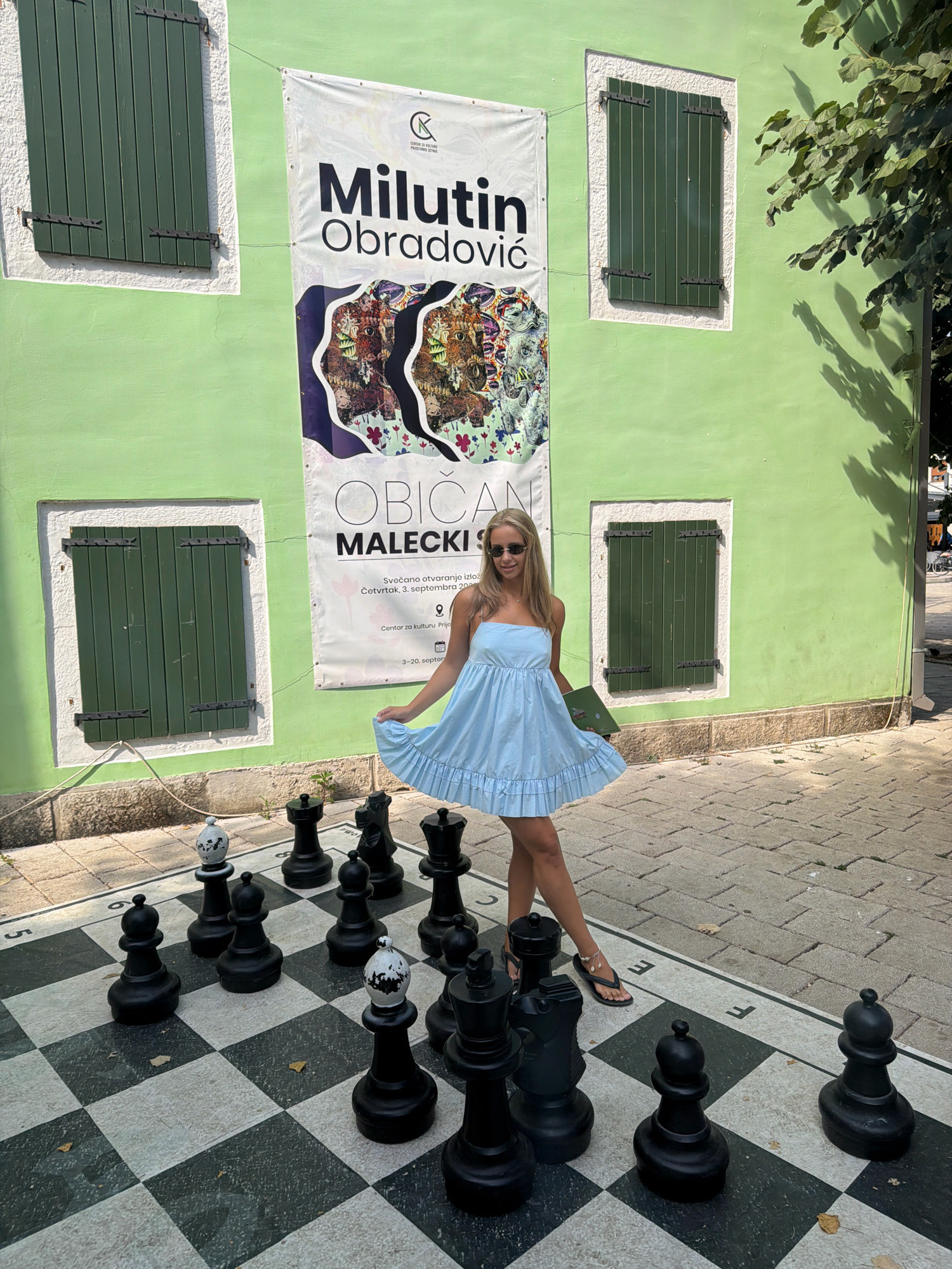 In front of a lime-green building with dark green shutters, a young woman in a light blue sundress poses next to an oversized chessboard. The banner behind her, likely an exhibition announcement for "Milutin Obradović" and "Obični Malečki," hangs on the building. The woman is wearing sunglasses and flip-flops. The chessboard, set up outdoors on a paved area, features large black and white pieces, including kings, queens, and pawns, some of which have white tops with black splotches. The scene is set on a sunny day.