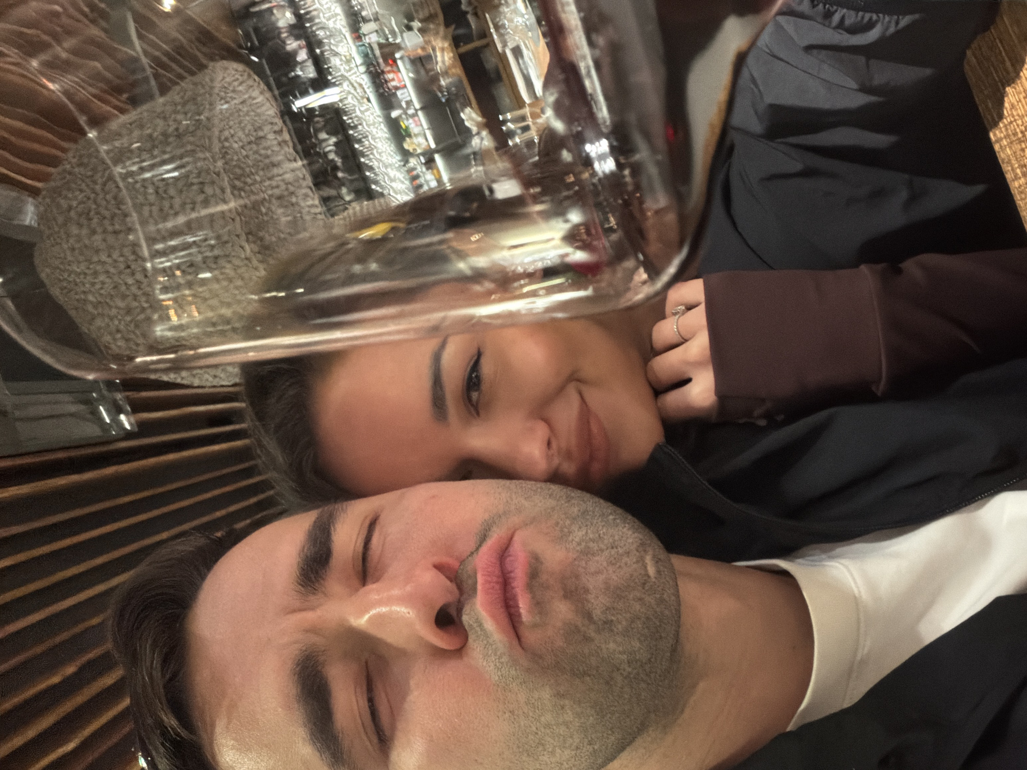 An intimate, close-up selfie-style portrait features two individuals, a man and a woman, positioned closely together. The image is taken at an angle, with the subjects appearing to lie down or lean back.
In the foreground, a man with light skin, short dark hair, and stubble covers much of his face. His eyes are closed or squinting, and his lips are slightly pursed, giving him a playful or contemplative expression. He wears a white t-shirt under a dark, possibly zipped-up, jacket.
Behind and slightly above the man, a woman with light skin and dark hair is visible, smiling gently. Her eyes are open, and she has visible eyeliner. Her left hand, adorned with a ring featuring a clear stone on her ring finger, is visible near her face. She wears a dark brown long-sleeved top, partially covered by a dark garment, possibly a jacket.
A prominent, large, clear, and highly reflective glass or plastic object occupies the upper portion of the image. This object reflects elements of the background, including shelves or displays filled with numerous items, possibly bottles or glassware, suggesting an environment like a bar or a well-stocked room. Behind the man's head, there is a patterned surface, resembling wooden slats or paneling.
The overall scene suggests an indoor setting, likely in the evening or at night, given the ambient lighting. The proximity and expressions of the individuals imply affection or intimacy, captured in a casual moment. No discernible text is visible in the image. The location context provided is Krasnoyarsk, Russia.