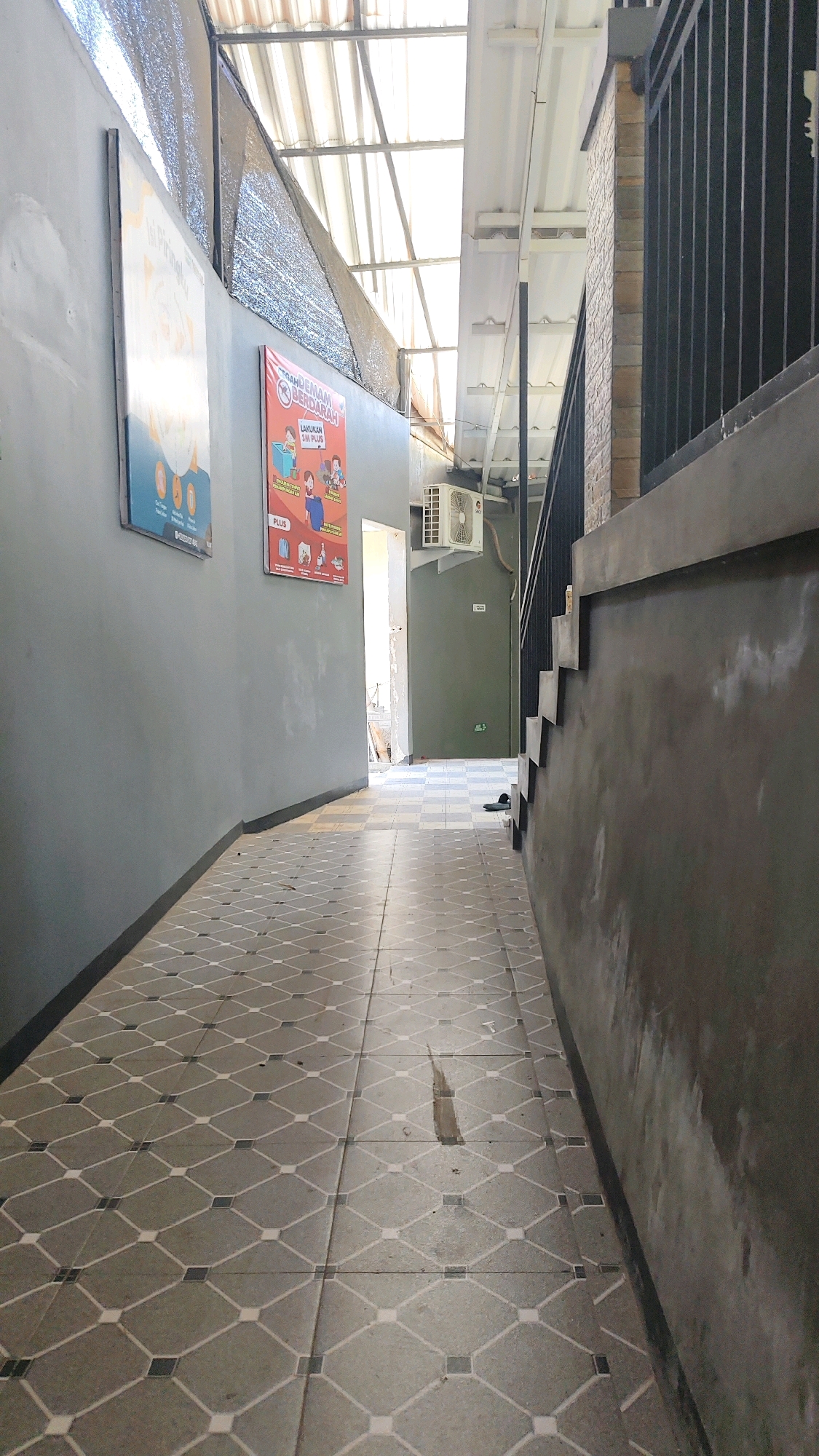 The image shows a narrow, covered walkway with patterned tiled flooring. The walkway is bordered by a grey wall on the left and a concrete structure with a black metal railing and brick accents on the right. Above, a translucent corrugated roof allows sunlight to filter through. Two posters are affixed to the left wall. One poster, on the left, is mostly white with some circular graphics. The second poster, to its right, is red and features text and images, including the words "DEMAM BERDARAH" and "LAKUKAN 3M PLUS". An air conditioning unit is mounted on the wall on the right side of the walkway, near the entrance to a darker hallway. A pair of black flip-flops are visible on the tiled floor in the distance. The scene suggests an outdoor or semi-outdoor area, possibly a passage or corridor, in a location that experiences warm weather, given the presence of the air conditioning unit and the outdoor-like roofing. Based on the provided context, this scene is located in the city of Tellok, Indonesia.