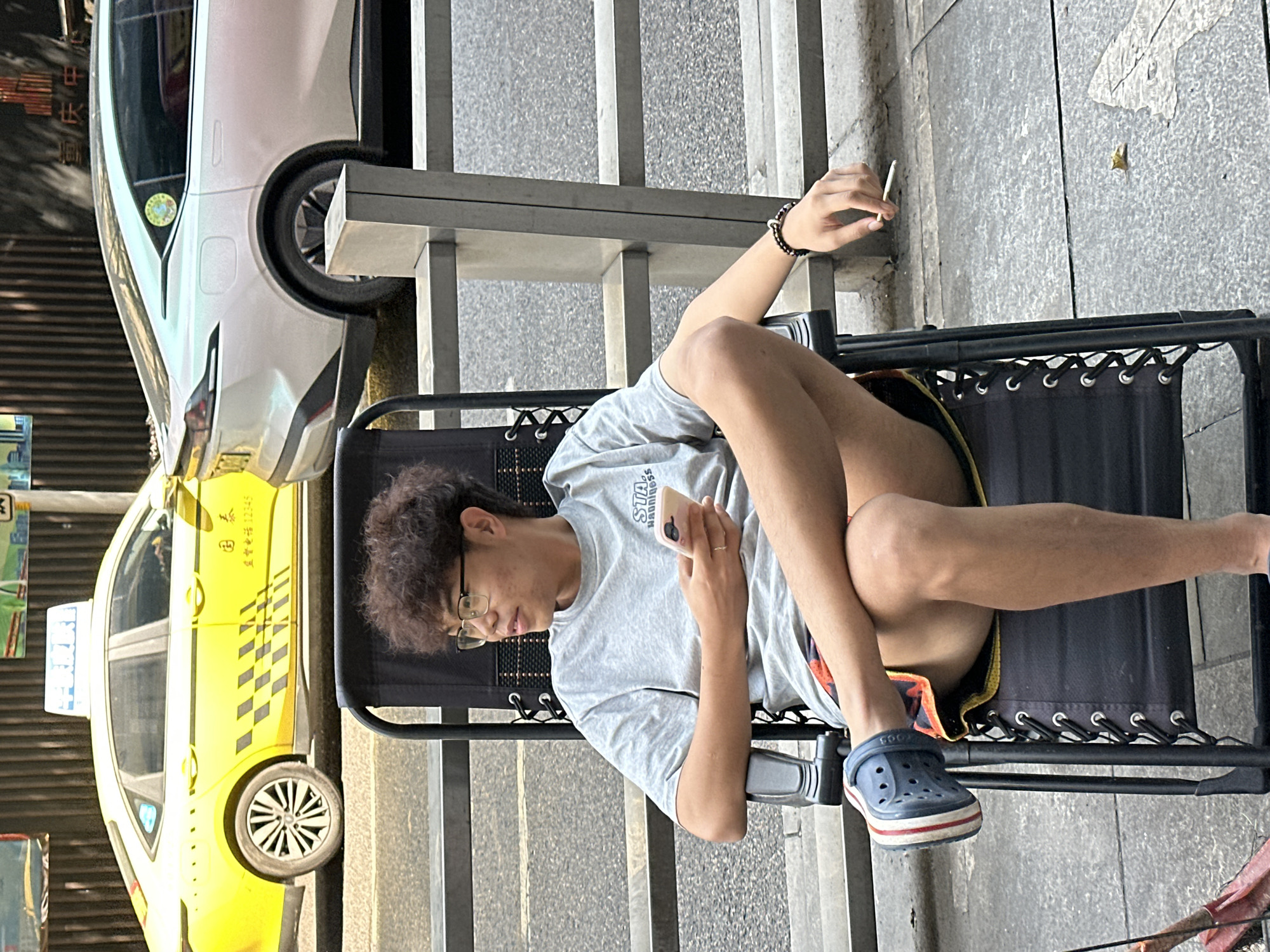 A man sits in a black folding chair on a sidewalk in Yuzhong District, China, near a taxi and a white vehicle. He holds a mobile phone in one hand and a lit cigarette in the other. He wears glasses, a gray t-shirt, and blue Crocs. The sidewalk is paved with gray stone. Stairs are visible behind the chair.