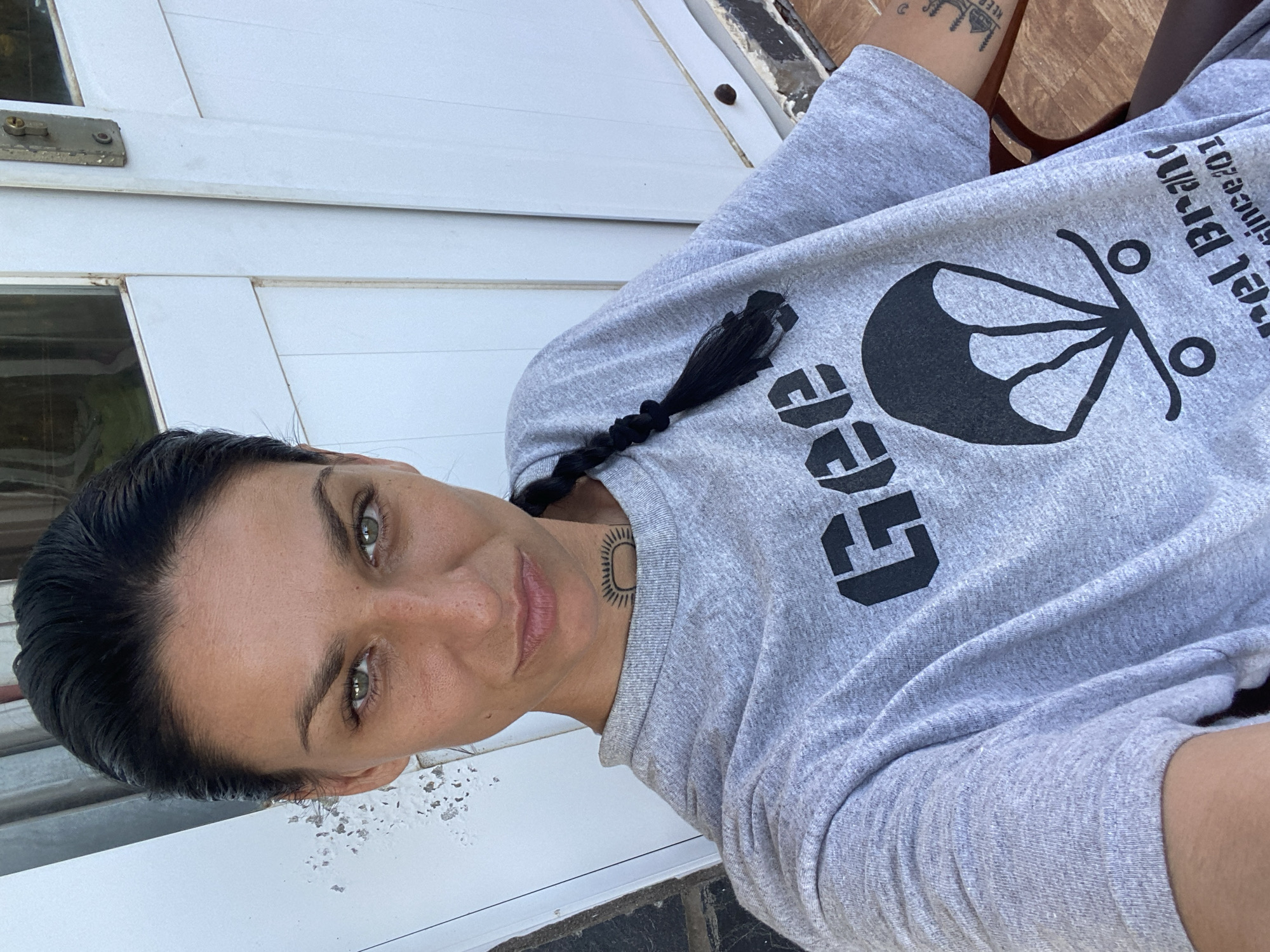 A woman with dark hair styled in a braid poses for a selfie. She is wearing a gray t-shirt with a black design on it, and has a tattoo on her neck. The background appears to be a white wall or door. The image may have been taken in Mũi Né, Vietnam.