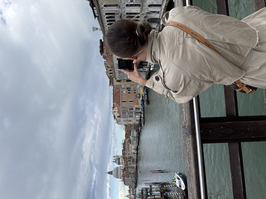A female individual is observed from behind, standing on the Ponte dell'Accademia in Venice, Italy, looking southwest along the Grand Canal. She wears a light beige trench coat and has dark hair styled in a bun. A brown leather cross-body bag with visible hardware hangs from her left shoulder. Both her hands hold a black smartphone in a vertical orientation, actively directed towards the panorama, suggesting photography or video capture.
In the foreground, a dark wooden bridge railing with a silver metal top rail is visible. Beyond the railing, the greenish-blue water of the Grand Canal stretches into the distance. Historic multi-story buildings with various window designs and terra cotta roofs line both banks of the canal. On the left side of the canal, in the mid-distance, the distinctive large dome and classical architecture of the Basilica di Santa Maria della Salute are clearly visible. A white and blue patrol-style boat, containing two figures, is moored to the left near a cluster of wooden poles. Numerous other smaller boats are docked along the canal's edges. The sky above is overcast with a blanket of grey clouds.