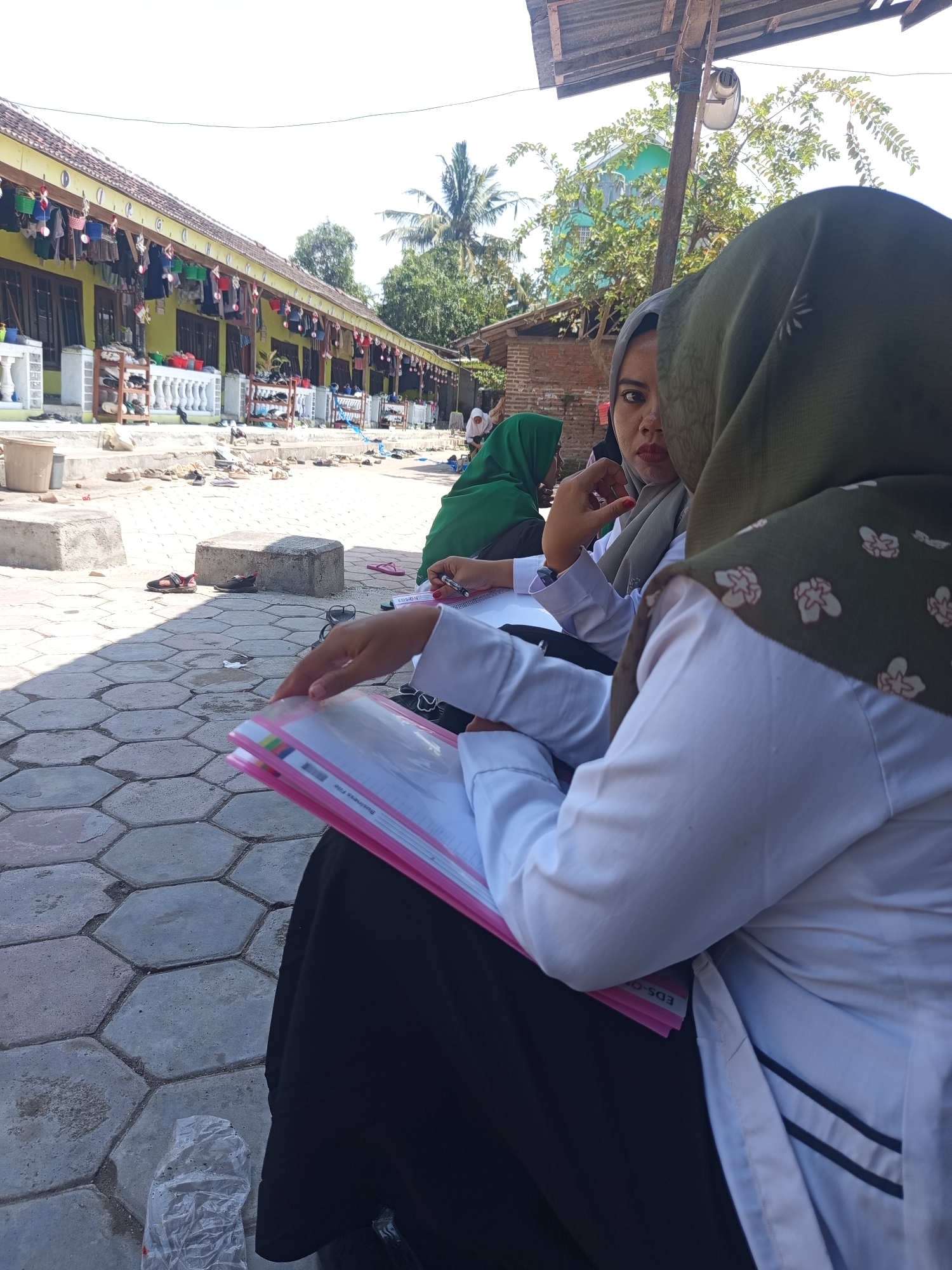 The image depicts an outdoor scene, likely in a residential or school setting in Lumutan, Indonesia. In the foreground, three individuals are seated, all wearing hijabs and modest clothing. One individual, facing partially towards the camera, is wearing a grey hijab and a white long-sleeved shirt. Another, whose back is to the camera, is wearing a dark green hijab with a floral pattern and a white long-sleeved shirt. A third individual, also with their back to the camera, wears a green hijab and a white long-sleeved shirt. One of the individuals in the foreground is holding a pink binder with documents.
The background reveals a row of yellow buildings with red-tiled roofs, possibly dormitories or classrooms. Clothes are hanging on display along the front of these buildings. Shoes are neatly arranged on shelves outside the buildings, suggesting a communal living or educational environment. The ground is paved with hexagonal interlocking concrete tiles. In the middle ground, there are concrete blocks and a trash can. Trees are visible in the background, with palm trees suggesting a tropical climate. The overall impression is one of an organized and functional community space.
