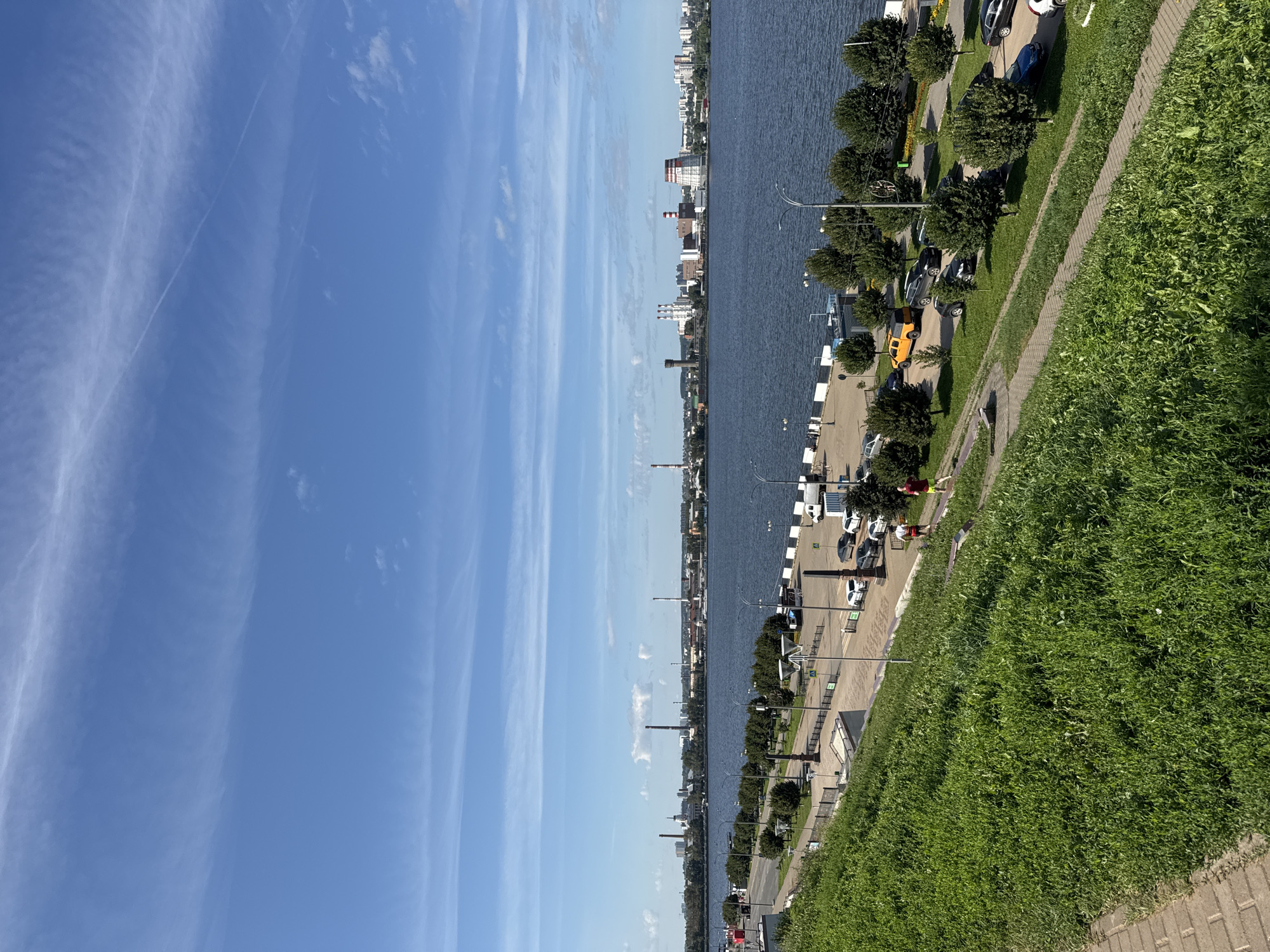 This image depicts a panoramic view of the Izh River embankment in Izhevsk, Russia. The foreground is dominated by a steep, grassy slope with a paved walkway winding down it, leading to a paved embankment area. On the embankment, there is a road with parked cars and people walking. The Izh River stretches across the midground, with a cityscape visible on the opposite bank. This cityscape features various industrial buildings, including smokestacks and a prominent cooling tower, as well as residential buildings. The sky above is a clear blue with scattered white clouds. The scene appears to be set on a sunny day.