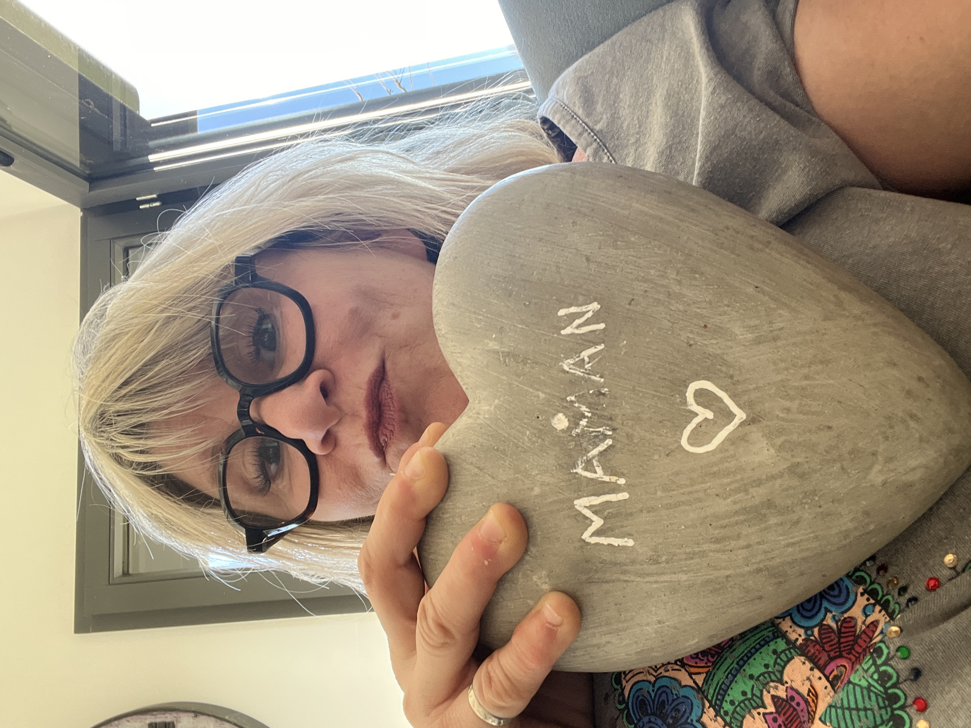A woman with blonde hair and glasses is holding a gray, heart-shaped stone with the word "Maman" and a heart symbol painted on it. She is wearing a gray shirt with a colorful floral pattern and rhinestones. The photo appears to be taken indoors, near a window. There is no indication of Chambœuf, France, in the image.