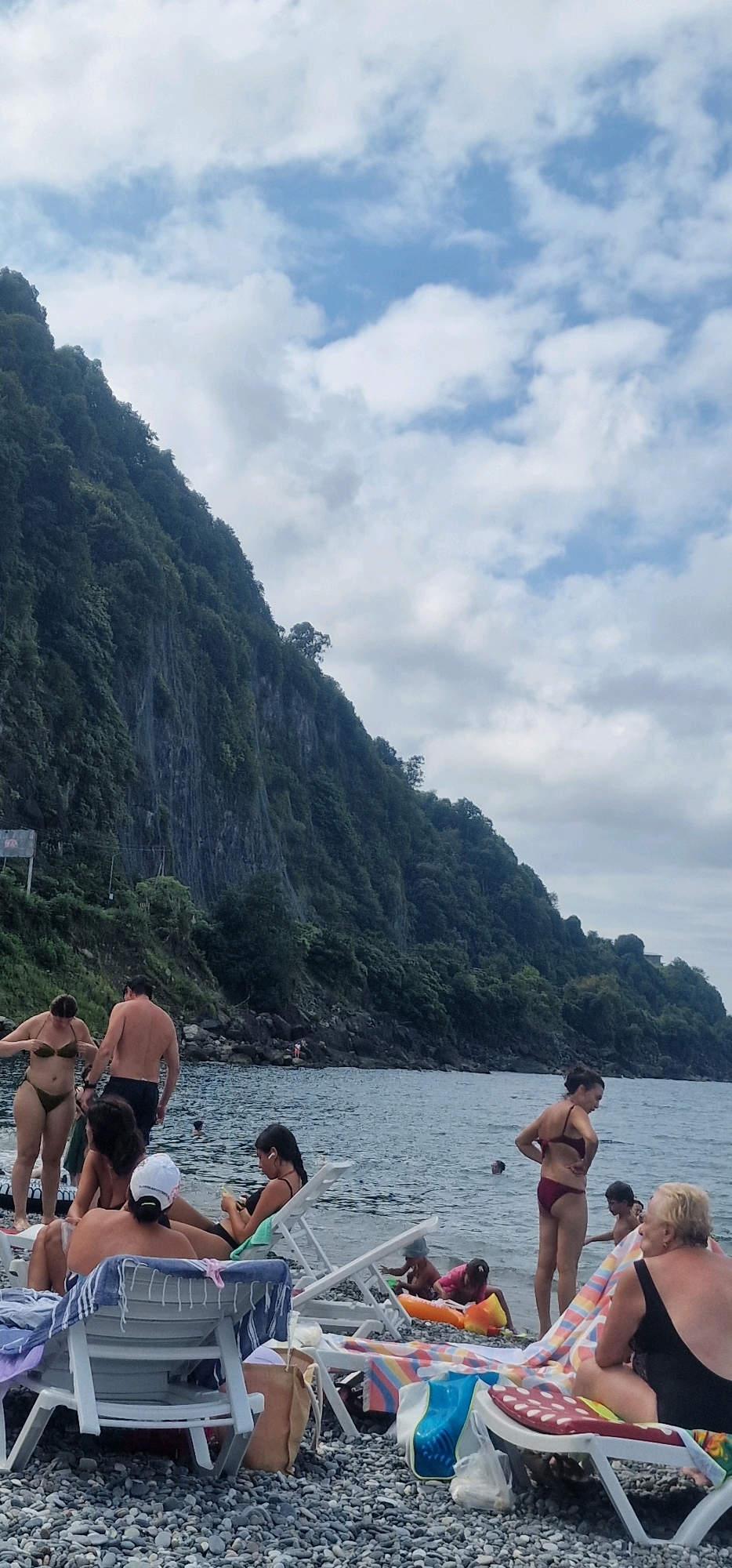 The image depicts a rocky beach scene in Batumi, Georgia, with a steep, verdant cliff face dominating the background. The sea is visible in the midground, with several people in and around the water. On the beach, there are multiple individuals relaxing on lounge chairs and sunbathing. Some are wearing swimwear. The sky is partly cloudy.