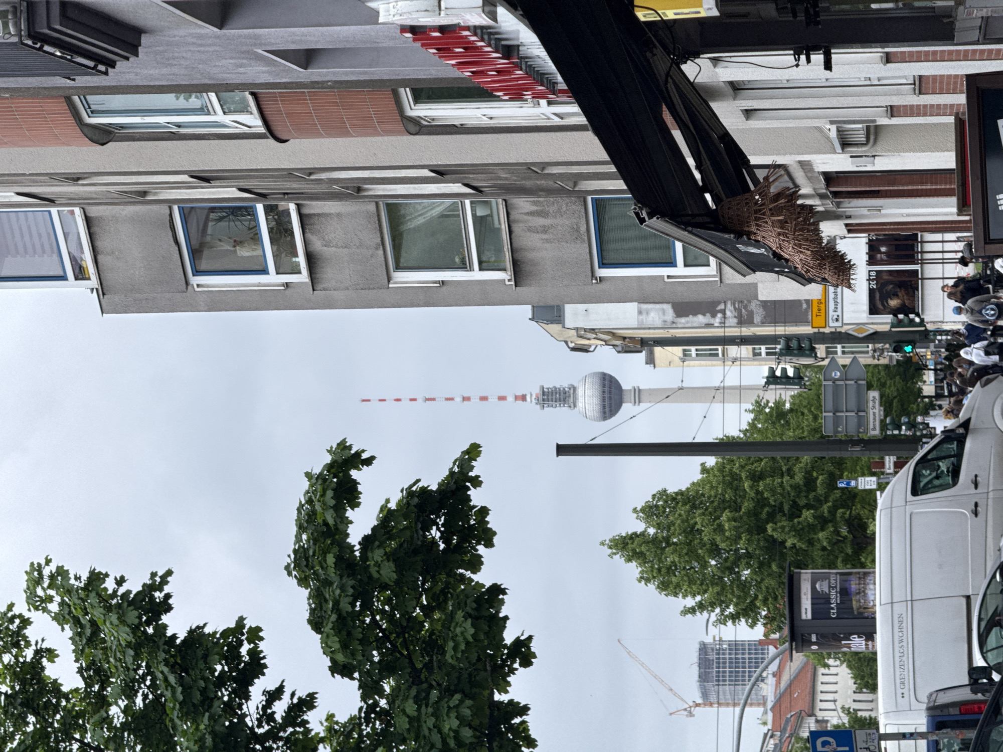 The image depicts a street scene in Berlin, Germany, dominated by the iconic Fernsehturm (TV Tower) in the distance. The foreground features dense green trees obscuring the lower part of the tower. Buildings line the street, with a mix of architectural styles visible. A white van is parked on the right side of the frame, partially obstructing the view. Street signs and traffic signals suggest a busy urban environment. The sky is overcast, creating a muted atmosphere. People and vehicles are visible on the street, indicating active daily life. An awning of a store or restaurant is seen at the top of the frame.