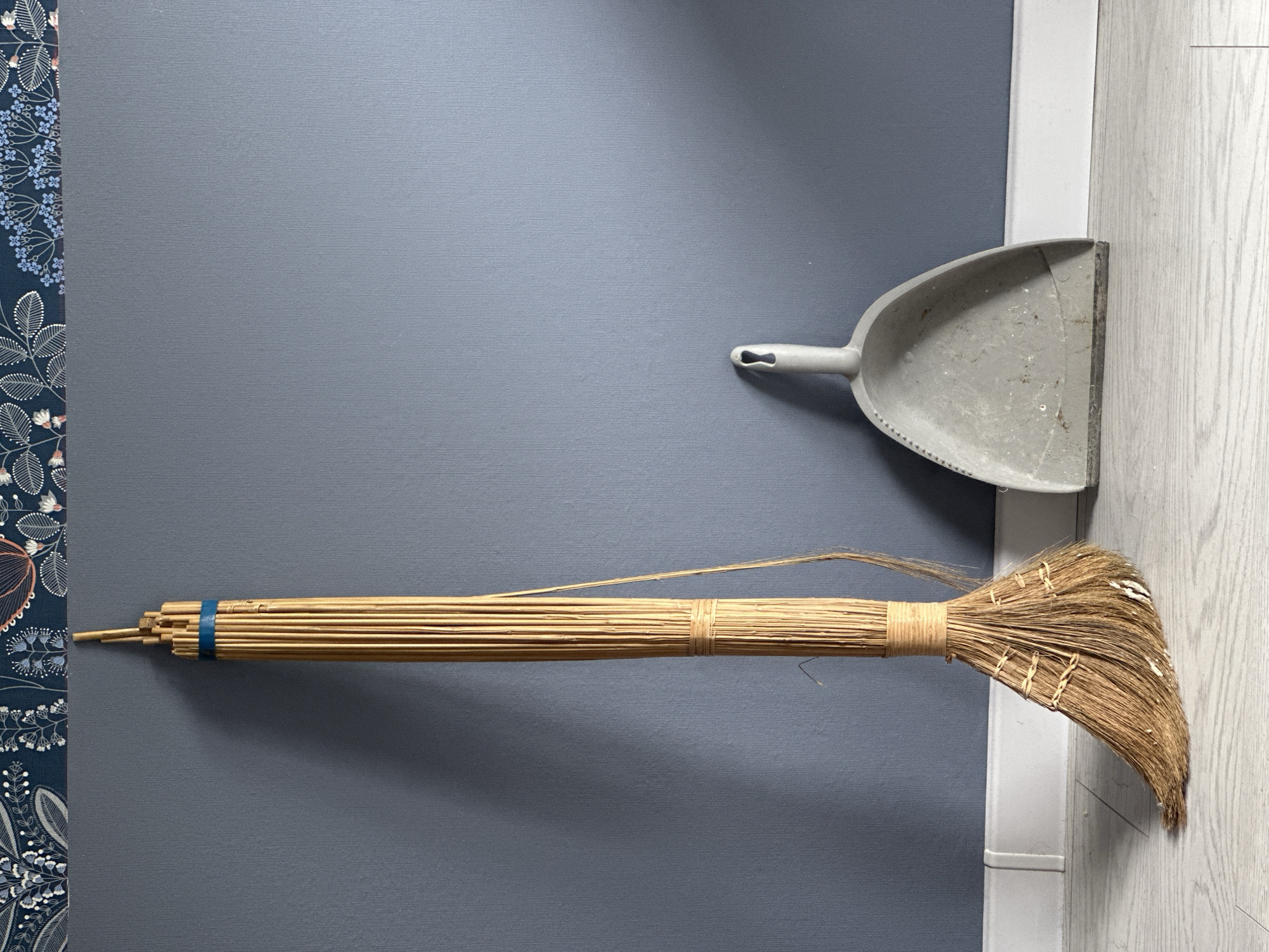 A broom and dustpan lie on a surface. The broom has a long, thin handle made of tightly bound straw or reeds, secured with blue tape at one end and twine in the middle. The broom's head consists of fanned-out, coarse fibers, possibly palm or similar material, also bound with twine. The dustpan, made of gray plastic, rests nearby. The background is a gray wall with a floral patterned border on the left and a white baseboard and light wood flooring on the right. There is no indication in the image of this being taken in Stavropol, Russia.