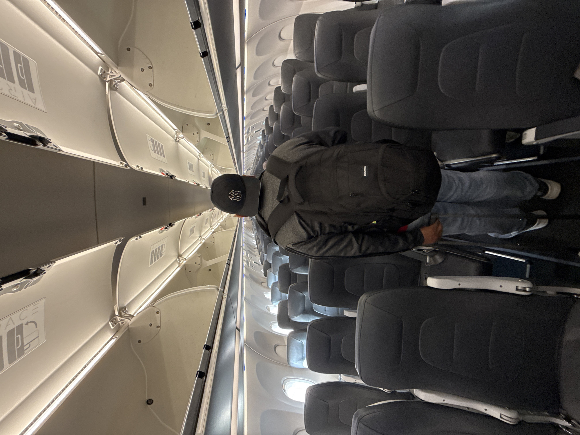 A man with a black backpack and a baseball cap is standing in the aisle of an airplane. The aisle is narrow and lined with rows of seats with gray upholstery. Overhead compartments run along the length of the cabin, illuminated by LED strip lights. The plane appears to be empty save for the man.
