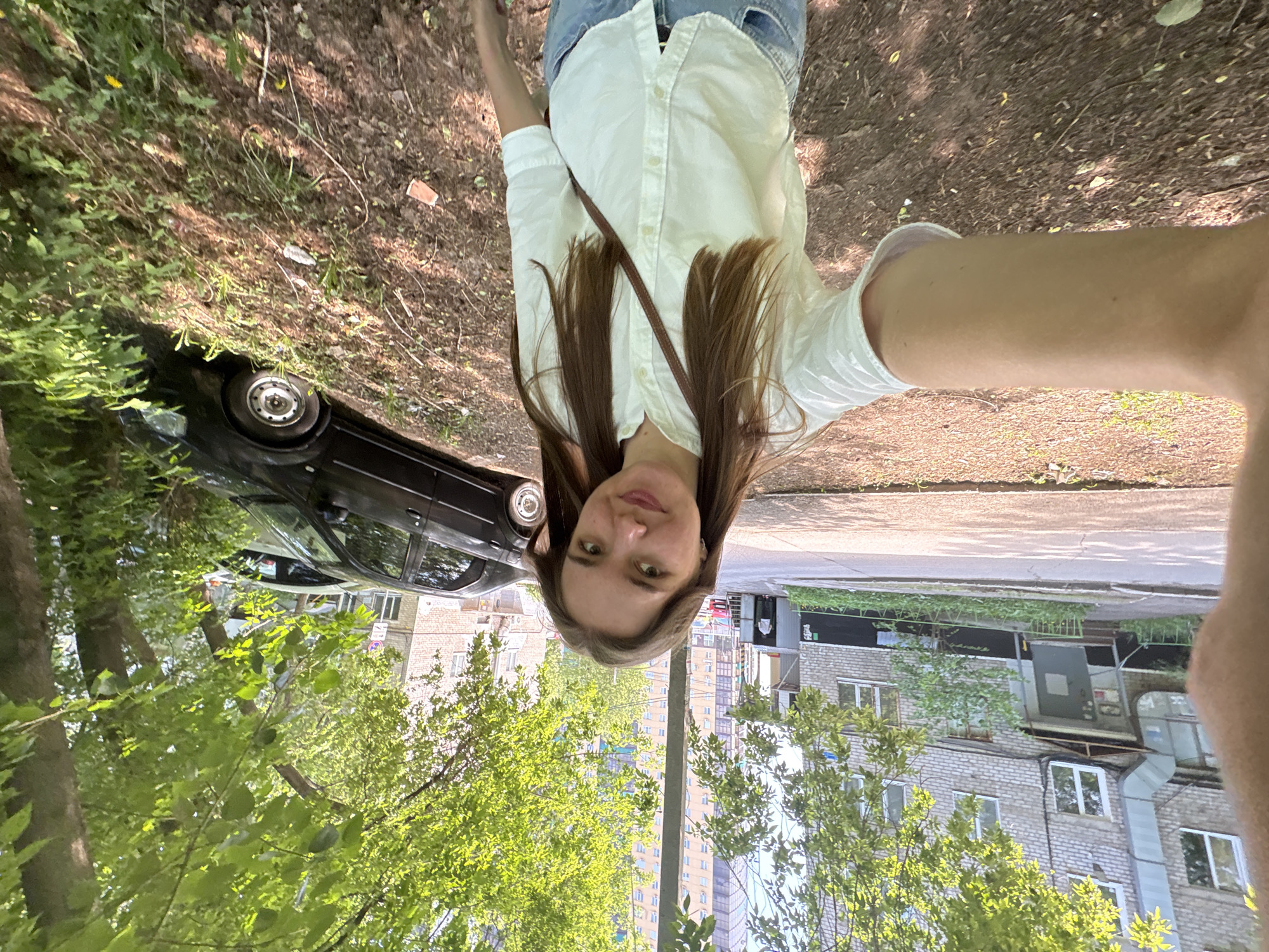 A selfie photograph shows a young woman in Perm, Russia. She stands with her upper body and head oriented vertically, wearing a light-colored button-down shirt and jeans. A brown strap crosses her chest. Behind her, the scene transitions from a roadside with dirt and vegetation to an urban environment with buildings and trees. A black car is parked to the left. Buildings are visible in the background. The location is Perm, Russia.