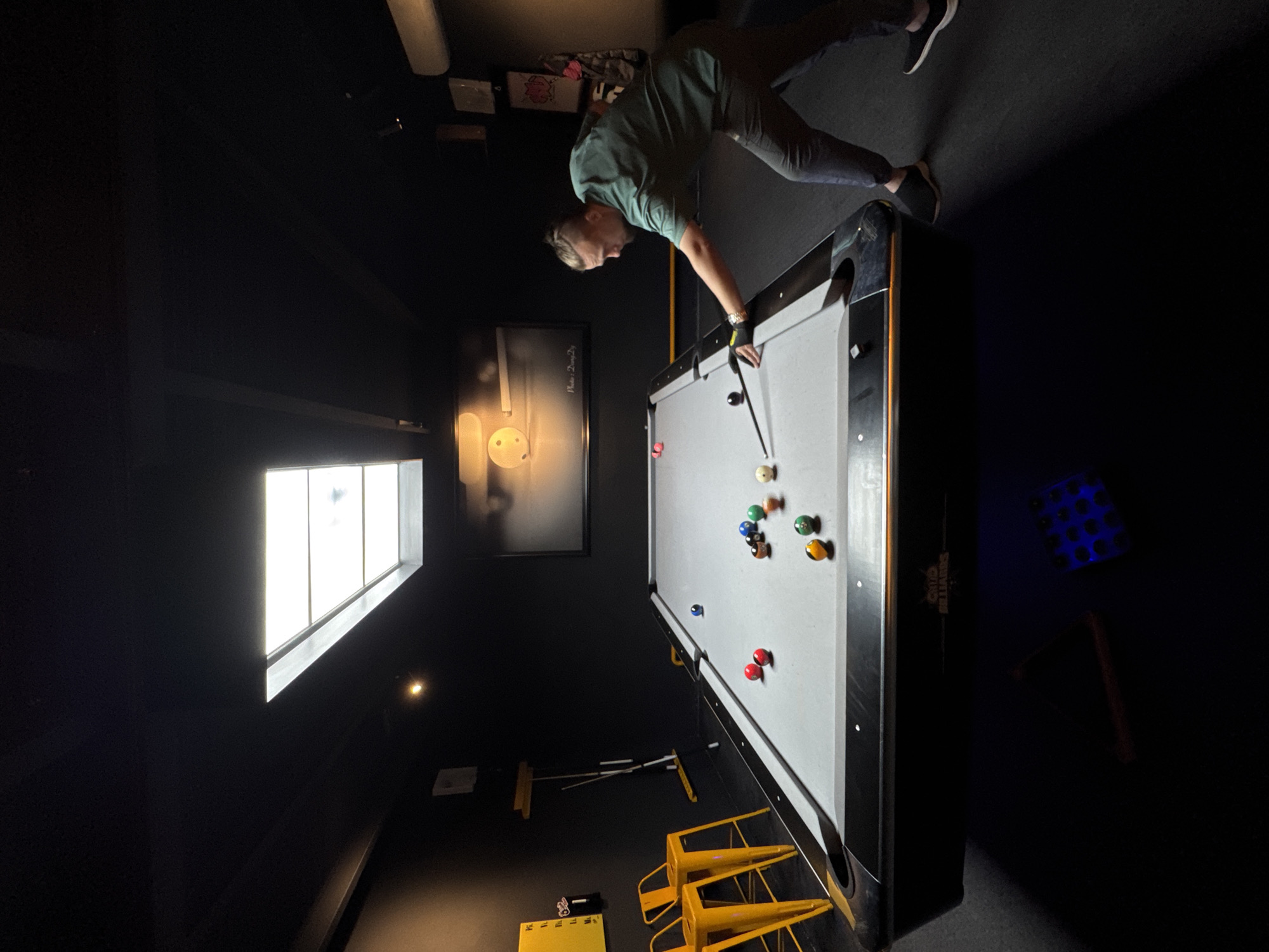 A man plays pool in a darkened room. He wears a green shirt and gray pants and leans over a pool table with a gray felt surface. Pool balls are scattered on the table. The room is dimly lit, with a window providing some light on the left. A framed photograph hangs on the wall above the pool table. Two yellow stools sit near the base of the pool table. The location is Phường Xuân Hương - Đà Lạt, Vietnam.