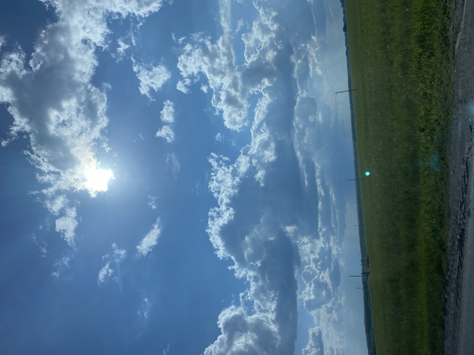 The image depicts a vast, open green field under a bright, cloudy sky. The sun is partially obscured by fluffy white clouds, casting a bright glow and creating a lens flare effect. The field appears to be a grassy meadow with scattered wildflowers and is bordered by a darker foreground, possibly a road or dirt path. In the distance, a line of trees forms the horizon, and several utility poles are visible scattered across the field. There are no people or animals present in the scene. The overall impression is of a rural landscape on a sunny day. The location is identified as the city of Tyumen, Russia.