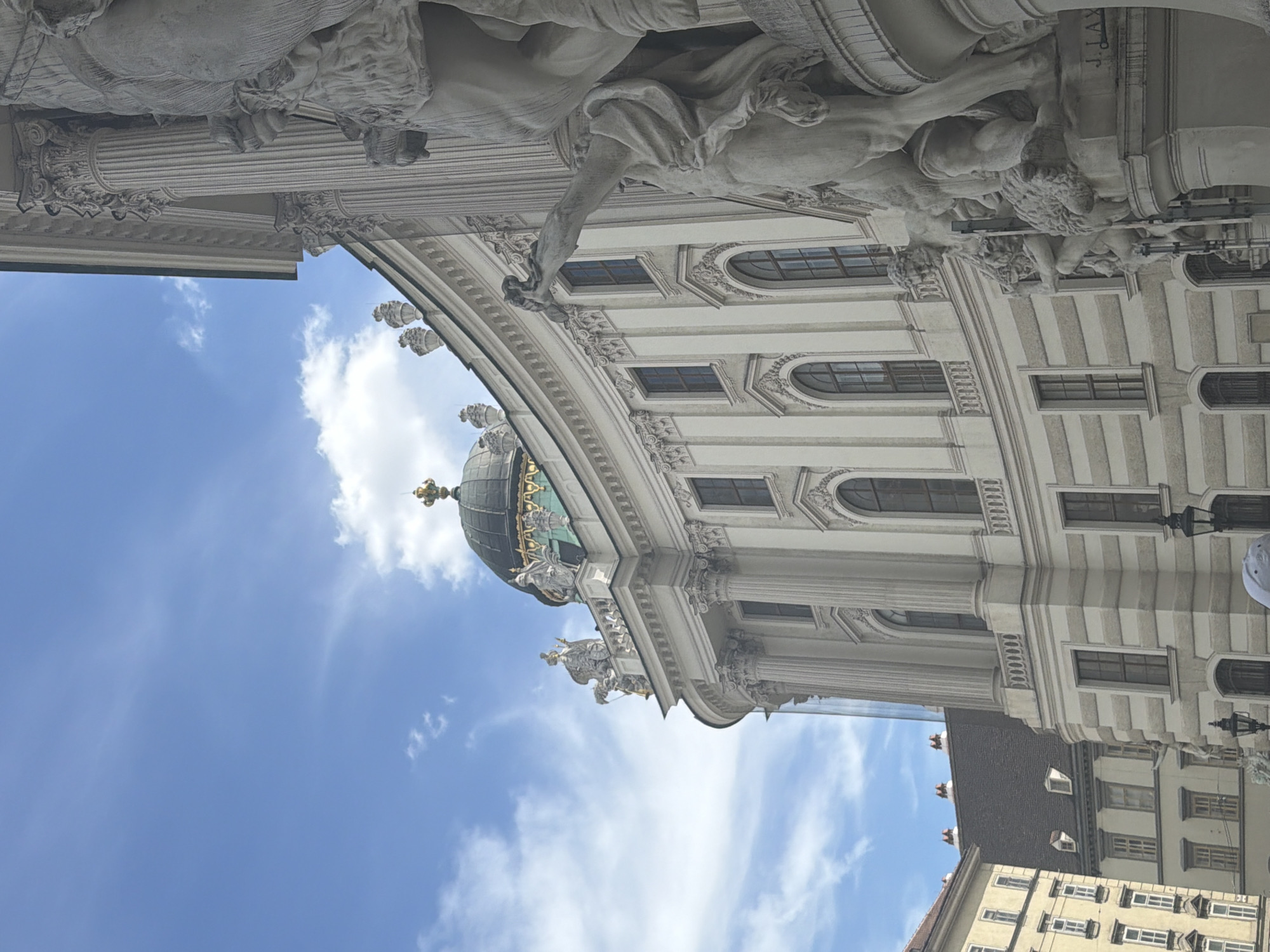 This is a photograph taken from a low angle, looking up towards a grand architectural structure against a bright blue sky with scattered white clouds. The building features ornate classical elements, including columns, elaborate carvings, and arched windows. A prominent dome, topped with a golden spire, is visible on the right side of the frame. Sculptural figures are integrated into the facade, with one large, detailed sculpture dominating the upper portion of the image. The architecture suggests a historical or religious building, consistent with the context of Vienna, Austria. The lighting indicates it is daytime, likely late morning or afternoon, with bright sunshine illuminating the scene. There is no discernible activity or event taking place; the image appears to capture a static architectural view. No visible text is present in the photograph.