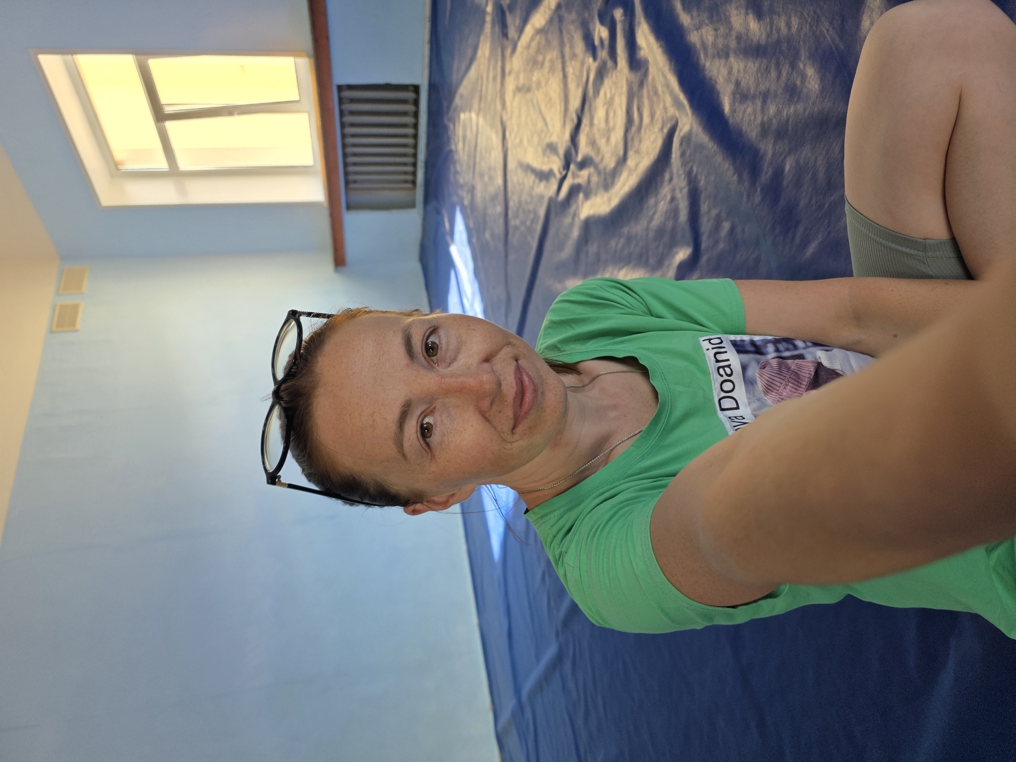 A woman with fair skin and freckles, wearing glasses on her head, a green t-shirt, and grey shorts, sits on a blue tarp indoors. She is holding her phone out to take a selfie. The background shows a light blue wall with two ventilation grates, a window with yellow light streaming through, a radiator, and a blue floor mat. The scene appears to be in a gymnasium or a similar recreational space. The location is indicated as the Chaikovsky City Hospital, Chaikovsky, Russia, suggesting this might be an area within the hospital premises.