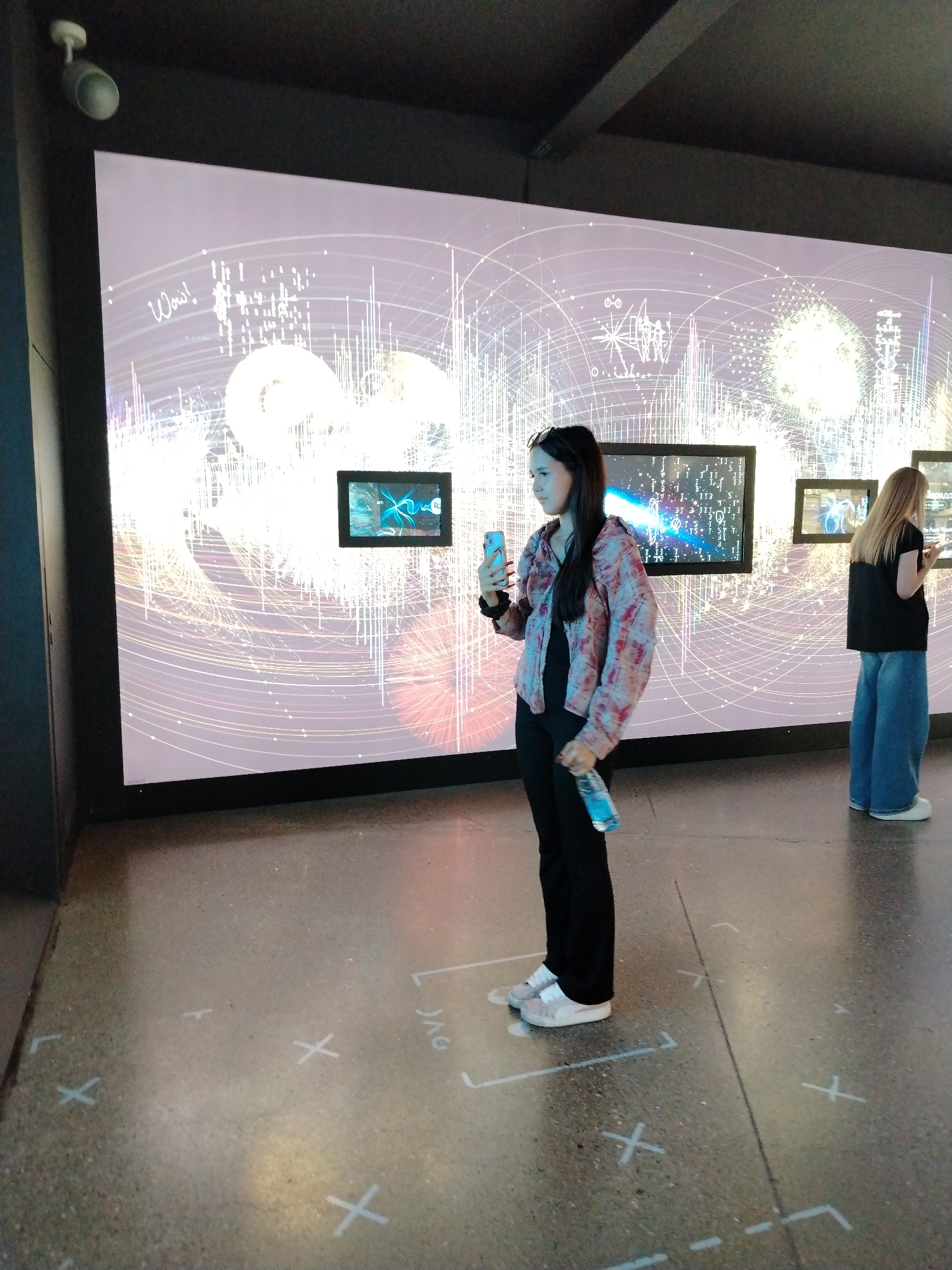 In Moscow, Russia, at the Museum of Cosmonautics, a young woman stands in front of a large digital display. She is holding a smartphone in her right hand and a clear plastic bottle of water in her left. She is wearing a black shirt underneath a patterned, open-front jacket and black pants, with white sneakers. A second woman, with blonde hair and wearing a black t-shirt and wide-leg blue jeans, stands further to the right, also looking at the display. The digital display shows a complex, abstract pattern of lines and geometric shapes in shades of white and pink, with what appears to be the word "Wow!" written in cursive on the left side. Several smaller screens are embedded within the larger display, showing animated images. The floor is a polished concrete surface with white markings, including crosses and lines that appear to denote designated viewing areas.