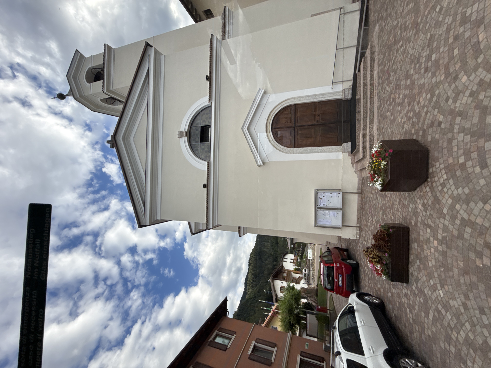 The image shows a street scene in the Italian town of Cavedago. In the foreground, there are two cars parked on a cobblestone street. The car on the left is a white SUV, and the car on the right is a red hatchback. In the middle ground, there is a church with a tall bell tower. The church is painted a light yellow color and has a large wooden door. In front of the church, there are two large planters filled with flowers. In the background, there are several buildings, including a red-colored building to the left of the church, and a backdrop of forested mountains. The sky is partly cloudy with blue patches. A sign in Italian and German is visible at the top left of the image, indicating emergency exit and emergency contact information. The scene appears to be a quiet, picturesque town square.
