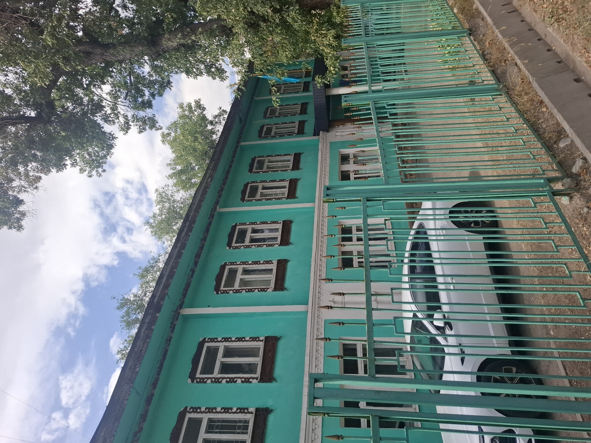 The image shows a turquoise-colored two-story building with white trim and decorative wooden window frames. The building appears to be an older structure. A light-colored modern sedan is parked in front of the building, partially visible behind a green metal fence. The fence has a geometric pattern with vertical bars and some decorative elements. Behind the building, trees are visible, and a blue and yellow flag is hanging from the side of the building. The ground in front of the fence is dirt with some dry grass and a concrete drainage ditch. The sky above is partly cloudy. The building is located on a street in Almaty, Kazakhstan, and appears to be a historical or culturally significant site, possibly a museum or a former administrative building, given the presence of the flag. The scene suggests a quiet daytime setting.