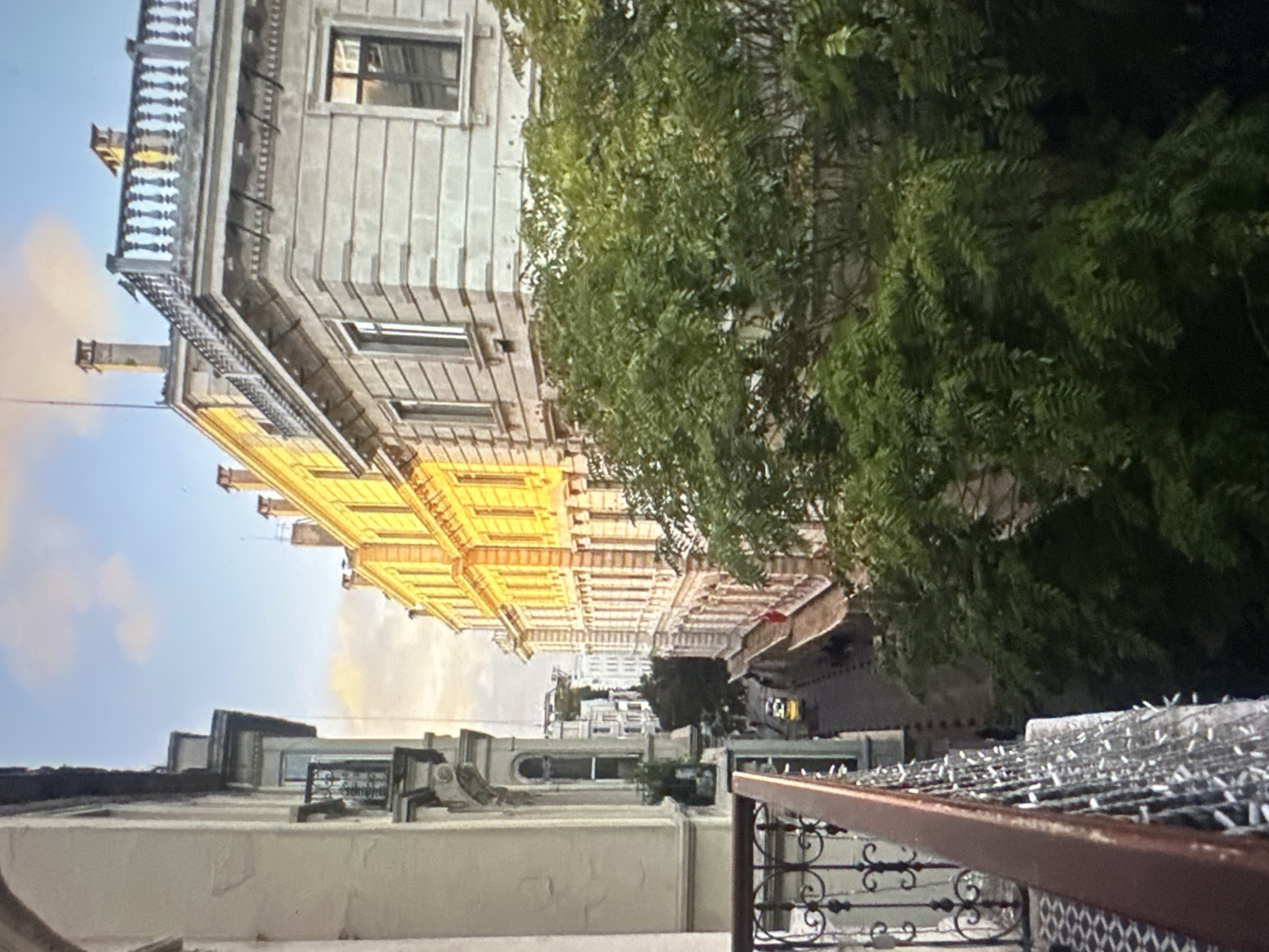 A vertical, high-angle shot captures a street view between two buildings, with lush green foliage obscuring much of the right side of the frame. The building on the left is a light-colored, ornate structure with a balcony in the foreground, featuring a dark brown railing and decorative black metalwork. The building on the right is a yellow, classical-style building with numerous windows and decorative stonework. The street below is narrow, with a yellow car and a few indistinct figures visible. The sky above is a light blue with scattered clouds, and sunlight illuminates the side of the yellow building, casting a warm glow. The overall scene depicts a European-style city street.