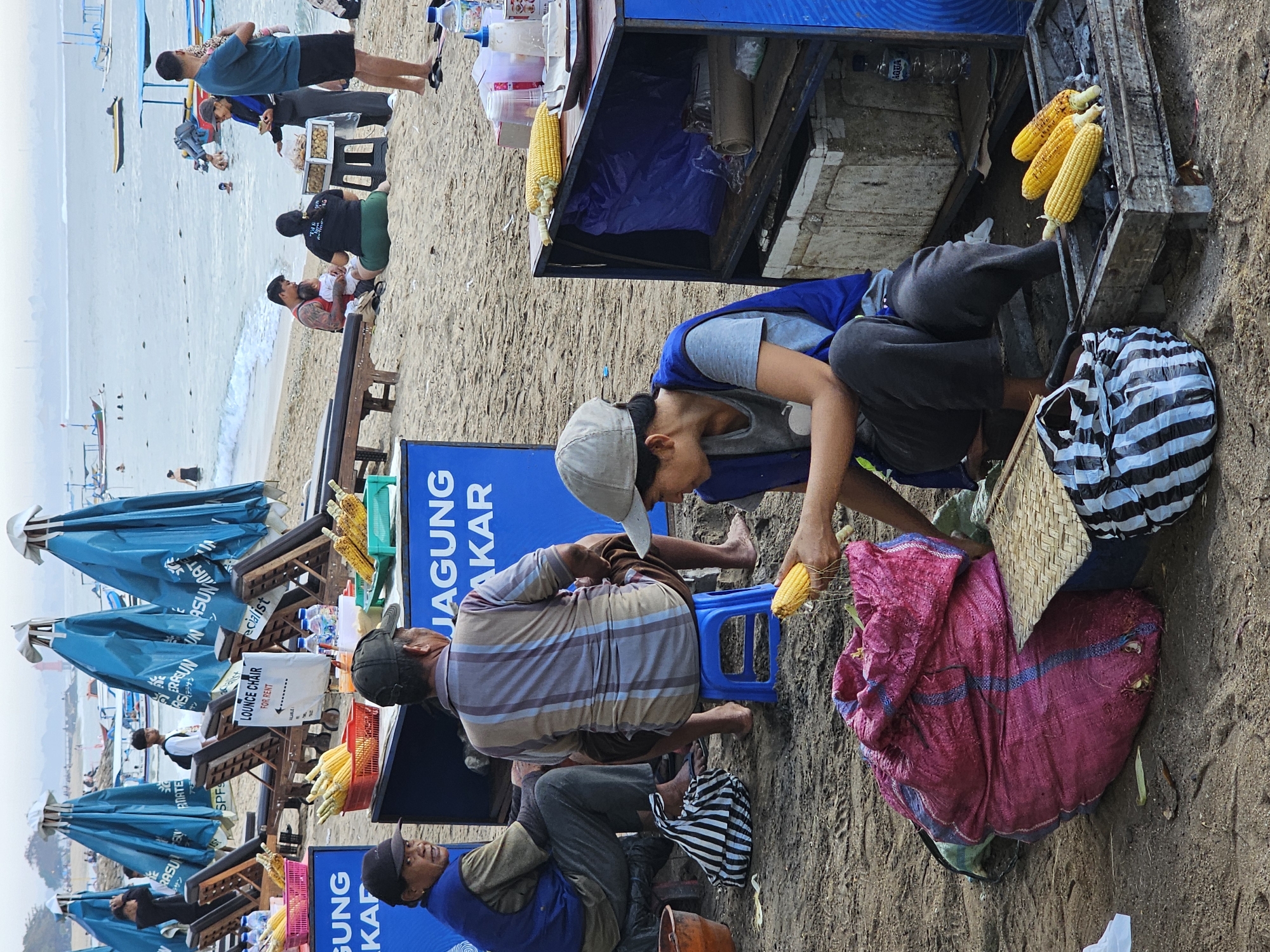 The image depicts a beach scene in Denpasar, Indonesia, likely in the vicinity of a popular tourist area given the presence of beach umbrellas, lounge chairs, and people engaged in leisure activities. The foreground features a beach vendor preparing corn. A woman, wearing a blue vest over a grey t-shirt and a grey cap, is seated and peeling a cob of corn. In front of her is a large, pink woven bag and a striped black and white cooler bag. To her right, a grill with three corn cobs is visible, suggesting they are being roasted. Behind the grill, a portable cart or stand holds various items, including bottles of water, condiments, and what appear to be more ears of corn.
To the left of the woman, two men are seated. One man wears a dark cap and a blue vest over a grey t-shirt, looking towards the viewer. The other man is seated on a small blue stool, wearing a striped shirt and dark shorts, facing away from the camera. Behind them are several blue beach umbrellas and wooden lounge chairs.
In the background, the ocean is visible with several boats anchored offshore. Other beachgoers can be seen in the water and on the sand. The overall atmosphere suggests a typical day at a busy Indonesian beach, with vendors providing services to tourists and locals. The sign on one of the umbrellas, partially visible, indicates "Lounge Chair For Rent." The blue cart in the middle ground has text that appears to be "JAGUNG BAKAR," which translates to "Grilled Corn" in Indonesian, further confirming the presence of a food vendor.