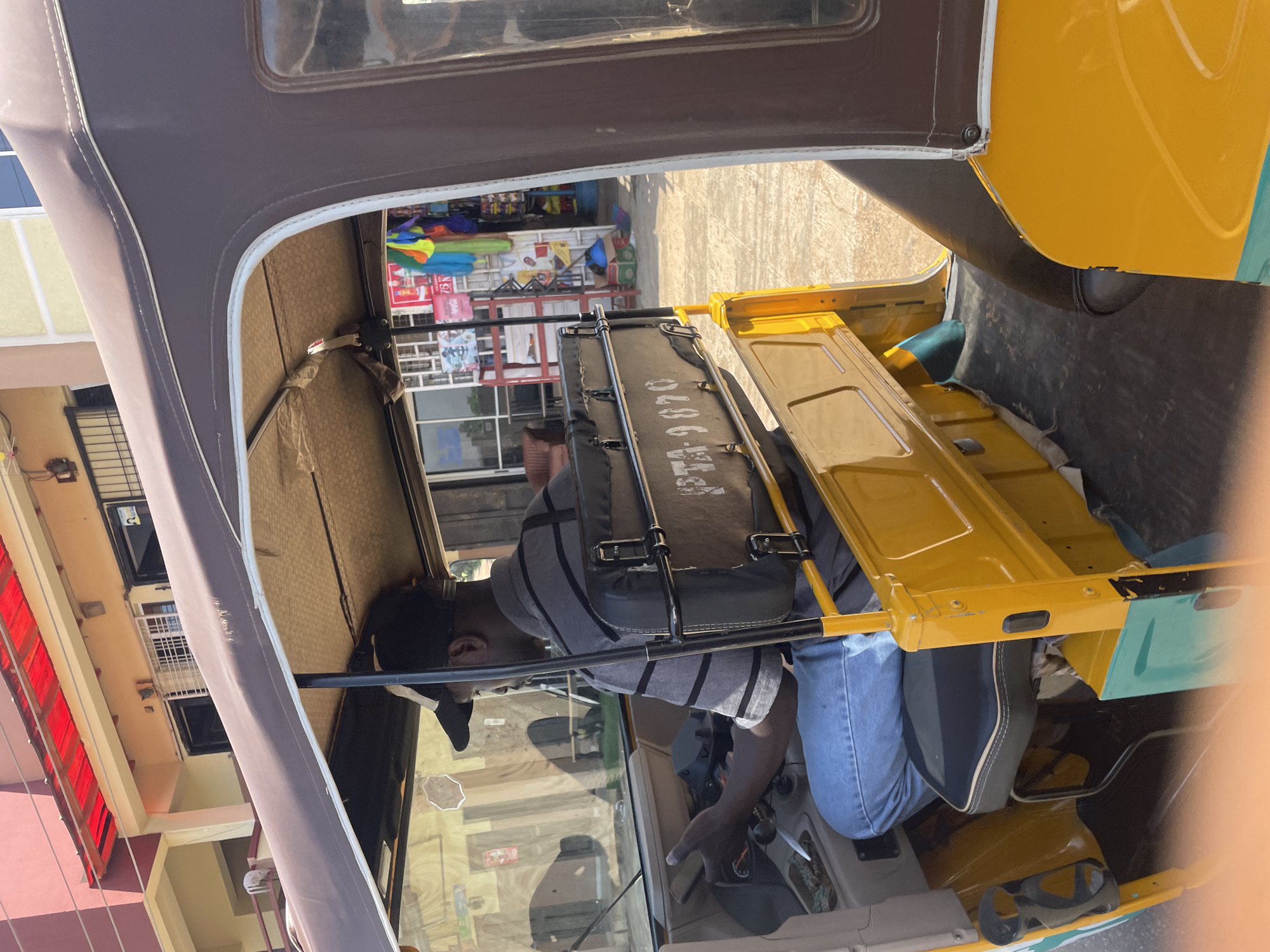 A man wearing a black baseball cap and a gray and black striped shirt is driving a yellow auto-rickshaw. He is wearing blue jeans and is holding the steering wheel. The auto-rickshaw has a brown fabric canopy and a black seat. The vehicle is parked in front of a building with a red awning. In the background, a store is visible with various items displayed on shelves. The ground is paved with a textured surface. The scene appears to be in an outdoor urban environment, possibly in Gura Top, Nigeria, given the context.