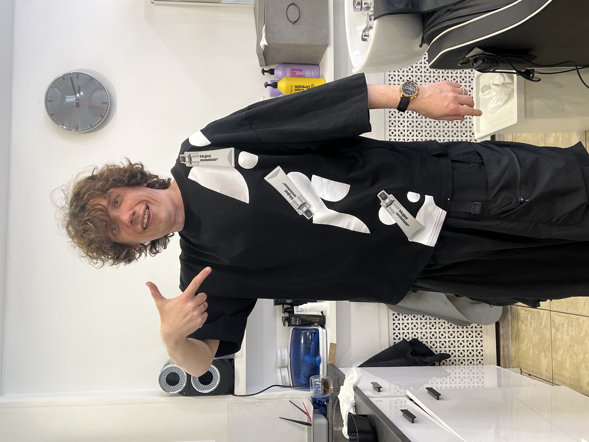 A man with curly brown hair, wearing a black t-shirt with white circles and tubes of "L'Oreal Majirel" hair color cream attached to it, is pointing with his left hand and smiling. He is standing in what appears to be a hair salon. The room is white with a dark gray clock on the wall. There are various hair products, a sink, and a black chair visible in the background. The person is wearing a black watch on his left wrist. The tubes are attached to the shirt near the shoulders and on the body of the shirt. There's a gray storage container with a hanging handle and various salon products like purple and yellow bottles. The salon appears clean and well-lit. Location and city information are NOT provided.