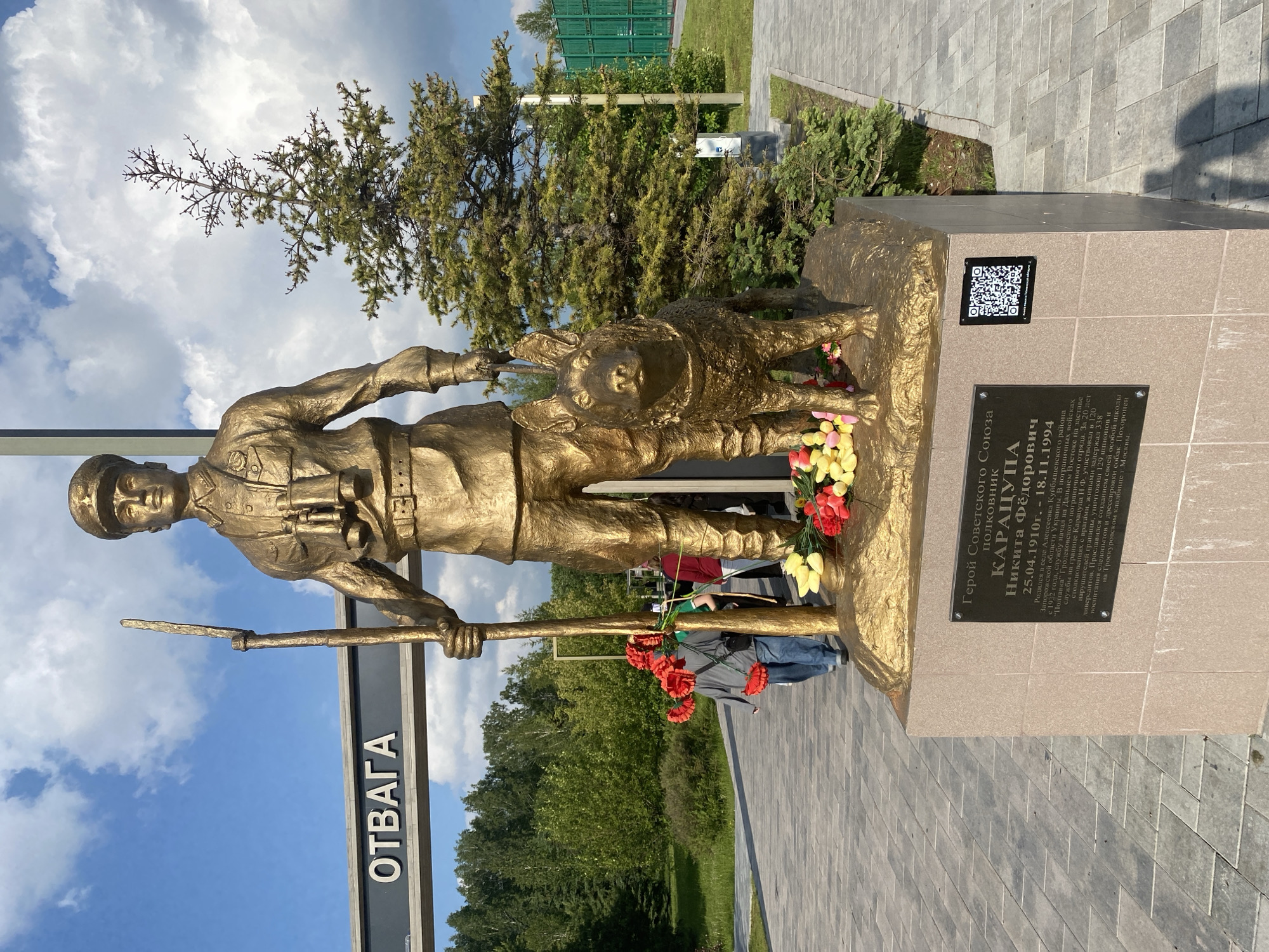 A bronze statue of Colonel Nikita Fedorovich Karatsupa, Hero of the Soviet Union, and his German Shepherd-type border guard dog is captured in a daytime, eye-level shot within the Park of Courage (Парк Отваги) in Tyumen, Russia.
The statue, rendered in a uniform bronze finish, depicts Karatsupa standing upright, wearing a military-style uniform with a cap, shirt, trousers, and boots. Binoculars are suspended from his neck, and a rifle or staff is held vertically in his right hand. His left hand holds a leash connected to the dog, which stands at his left side, facing forward. The entire sculpture rests on a rectangular pedestal constructed from light-colored stone.
Affixed to the front face of the pedestal is a dark rectangular plaque bearing white Cyrillic text that identifies the subject as "ГЕРОЙ СОВЕТСКОГО СОЮЗА полковник КАРАЦУПА НИКИТА ФЕДОРОВИЧ 25.04.1910. – 18.11.1994" (Hero of the Soviet Union Colonel Karatsupa Nikita Fedorovich 25.04.1910. – 18.11.1994). The plaque details his military service, including his role as a border guard, his capture of numerous border violators, saboteurs, and spies, his training of service dogs, and his burial in the Proskurovsky cemetery in Moscow. A black and white QR code on a small black plate is attached to the right side of the pedestal. At the base of the statue, on the pedestal, are multiple bouquets of artificial flowers, including red and yellow tulips and red poppies.
In the midground, to the left of the statue, a large grey horizontal sign with the white Cyrillic word "ОТВАГА" (COURAGE) marks an entrance or feature of the park. A paved pathway of light grey rectangular tiles extends from the foreground past the monument. Three individuals are partially visible near the base of the statue: one person in a grey jacket and blue jeans, carrying red flowers, walks away from the statue; another person in a green shirt is partially obscured behind the first; and a third person in a red top is further back.
The background consists of a dense line of green deciduous trees to the left, and a tall, slender conifer tree to the right, with additional evergreen shrubs at its base. A dark green metal fence or barrier is visible further to the right. The sky overhead is partly cloudy, with scattered white cumulus clouds. The scene is illuminated by natural daylight, casting shadows from the statue onto the paved pathway.