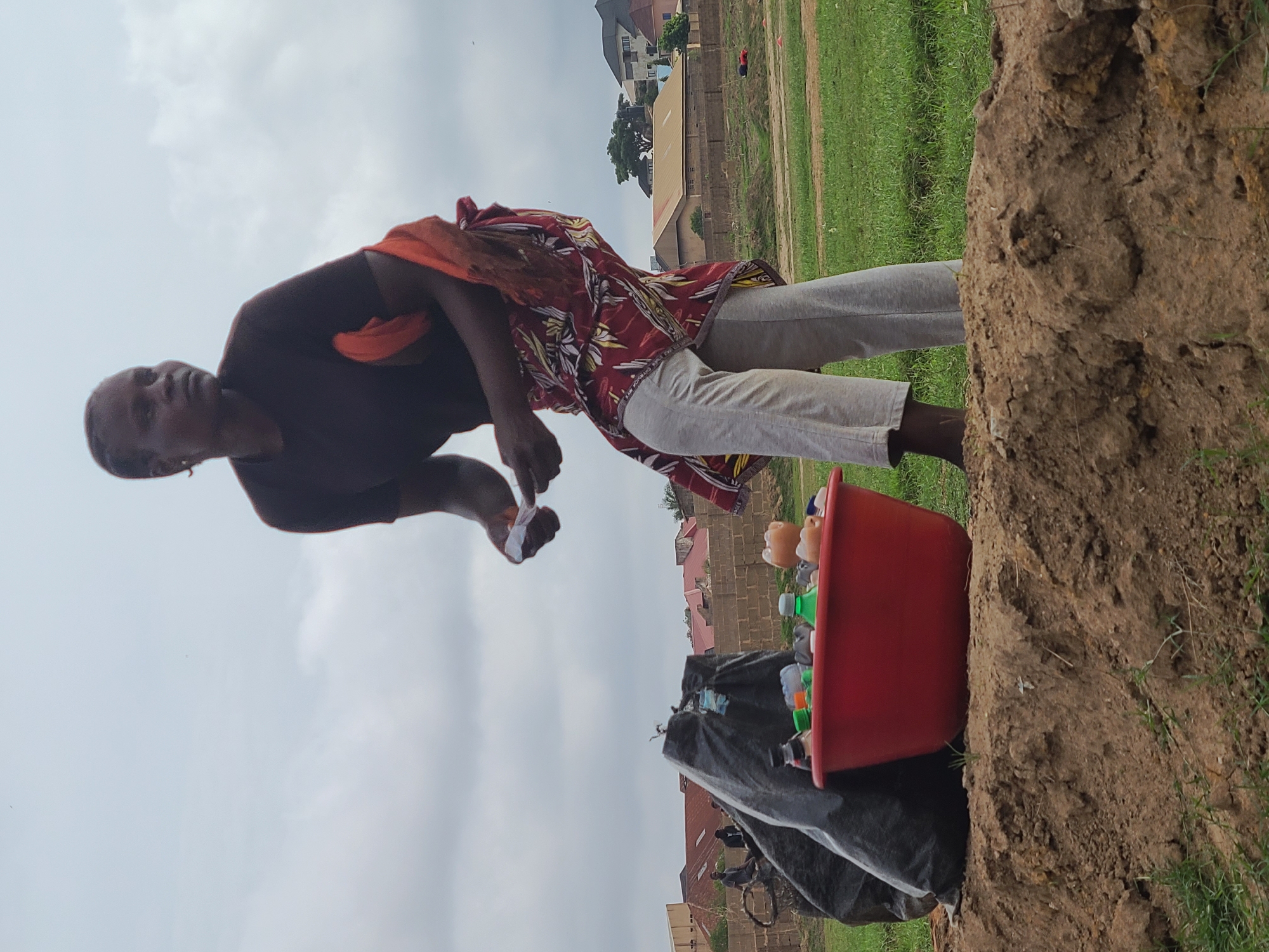 The image, captured in portrait orientation, features an adult individual standing outdoors next to a large mound of reddish-brown earth. The person, with dark skin, is wearing a dark short-sleeved top and light grey trousers. An orange fabric is draped over their left shoulder and neck. They are looking towards the camera with a neutral expression, holding a small white object in their hands, possibly in the process of sorting or inspecting it.
To the right of the individual, a red plastic basin is placed on top of the dirt mound. The basin is filled with numerous empty plastic bottles, some clear/brown and others green. Below the basin, a dark-colored bag or tarp rests against the mound, suggesting further collected materials.
The background reveals a landscape with green grass extending to the right. Beyond the mound and grass, several buildings with red roofs and brown walls are visible, indicating a populated area. The sky is uniformly bright and overcast, suggesting it is daytime. The scene conveys an impression of activity related to collection or recycling in a rural or semi-urban outdoor setting. No visible text is present in the image.