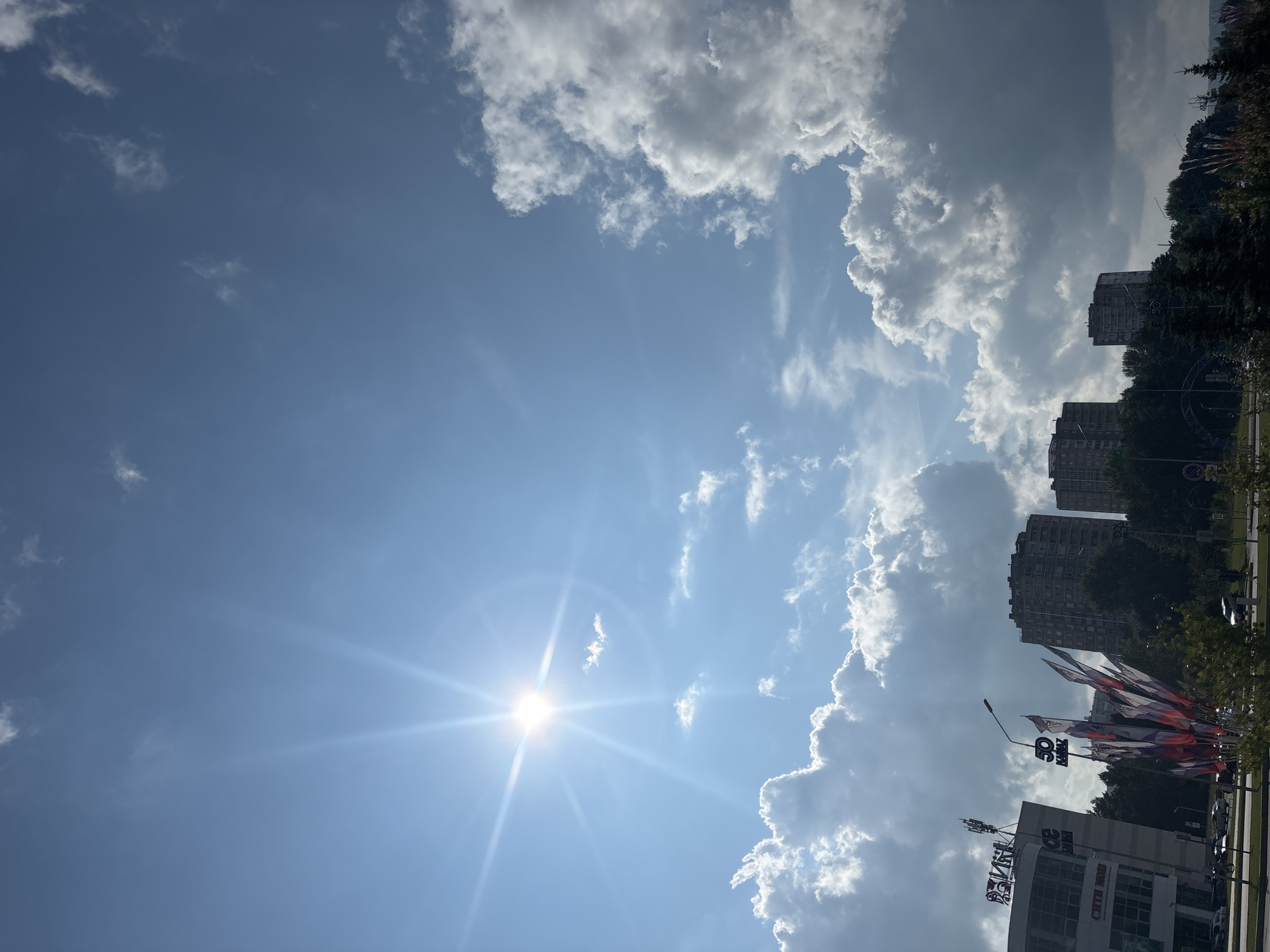 This image captures a bright, sunny day in Naberezhnye Chelny, Russia, with the sun prominently positioned in the upper left quadrant, casting a brilliant glare and visible rays. The sky is a clear blue, dotted with fluffy white clouds, some of which are partially silhouetted against the sun, creating a dramatic contrast. In the foreground, several tall apartment buildings are visible, nestled among lush green trees. To the left, a modern building with signage, possibly a shopping center, stands out, adorned with cellular antennas on its roof. A cluster of flags, displaying the Russian tricolor and other designs, are prominently displayed in front of the buildings, adding a festive or patriotic element to the scene. The lower part of the image shows a street with a few cars and what appears to be a road intersection, suggesting an urban setting. The overall atmosphere is one of a vibrant, modern city on a clear day.
