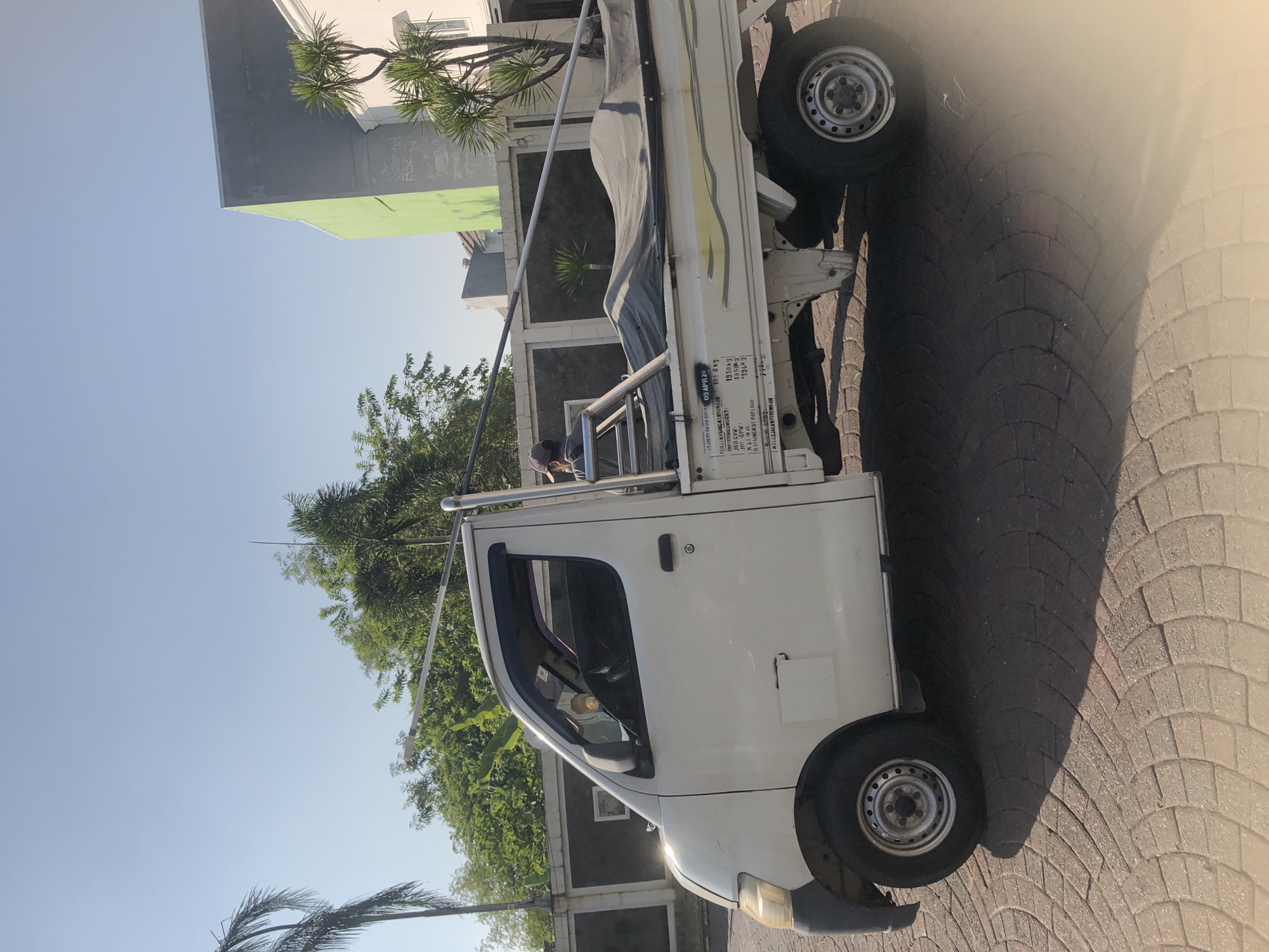 A white pickup truck is parked on a patterned brick surface. The truck has a black tarp covering its bed, and a person wearing a cap is visible in the driver's seat. Behind the truck, a low black wall with white accents runs horizontally, and beyond that, lush green trees and buildings are visible under a clear blue sky. A tall, thin palm tree stands to the left of the truck, and a green building is present to the right, behind the truck. The scene suggests a sunny day in a semi-urban or residential area of Boro, Indonesia.