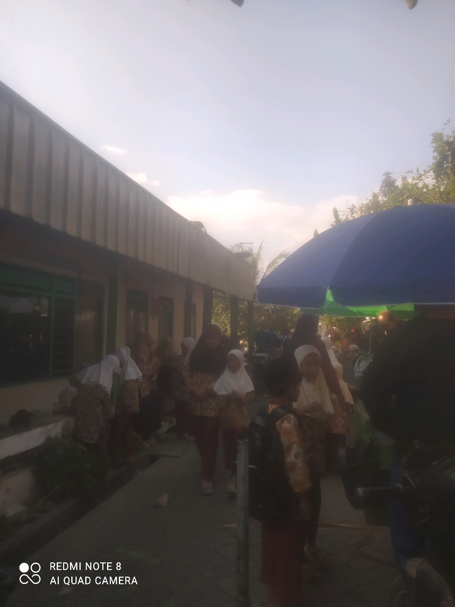 The image depicts a group of people, some of whom appear to be children, gathered outside a building. The setting is likely a street or pathway adjacent to a single-story building with a corrugated metal roof overhang. Several individuals are wearing headscarves and traditional patterned clothing. A large blue umbrella is visible, casting a shadow over some of the people. The sky is bright, suggesting daytime. The lower right corner of the image includes a watermark indicating "REDMI NOTE 8 AI QUAD CAMERA." The overall atmosphere is one of a casual gathering or activity in a semi-urban or rural environment. Based on the watermark and common mobile phone camera interfaces, this image was captured by a Redmi Note 8 phone. The location is identified as Wotan, Indonesia.