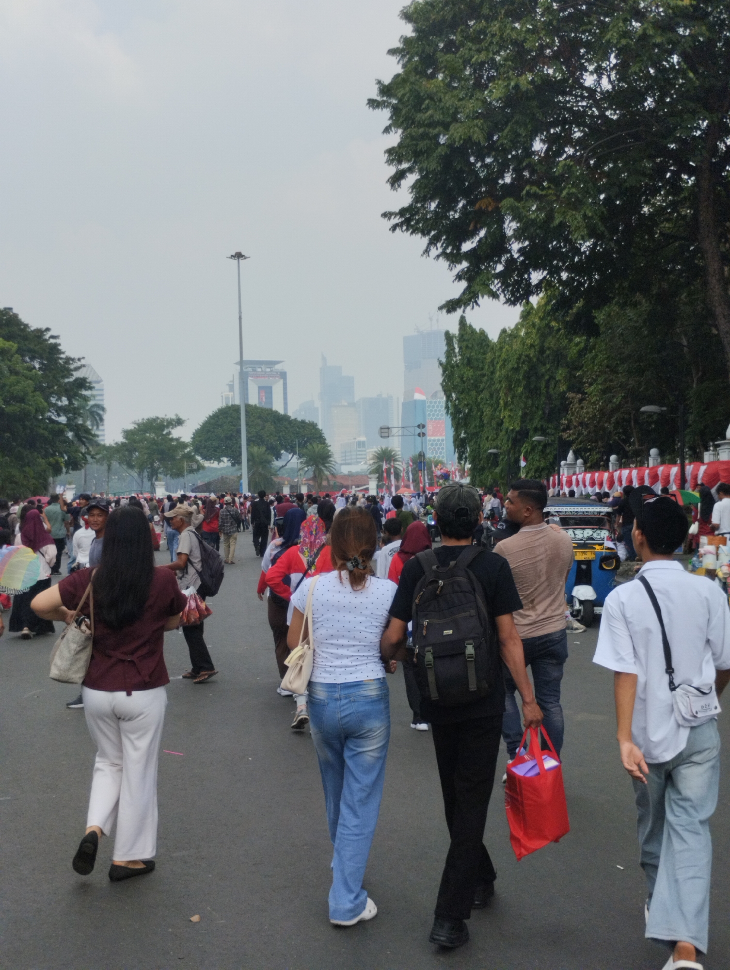 The image depicts a bustling street scene in Central Jakarta, Indonesia, possibly during a public event or festival, indicated by the large gathering of people and decorative elements. A crowd of individuals are walking along an asphalt road lined with trees and tall buildings in the background, suggesting an urban environment. In the foreground, several people are walking away from the camera. A woman in a maroon shirt and white pants is on the left, carrying a large beige bag. Next to her, a woman in a blue patterned shirt and blue jeans walks with her back to the camera. Further ahead and to the right, a man wearing a black shirt and backpack is walking, holding a bright red bag. To his right, another man in a light brown shirt and jeans is walking. Further down the road, a blue and white three-wheeled vehicle, possibly an auto-rickshaw, is visible. Red and white banners are displayed along the right side of the road, adding to the festive atmosphere. The sky is overcast, and the overall lighting suggests it is daytime.
