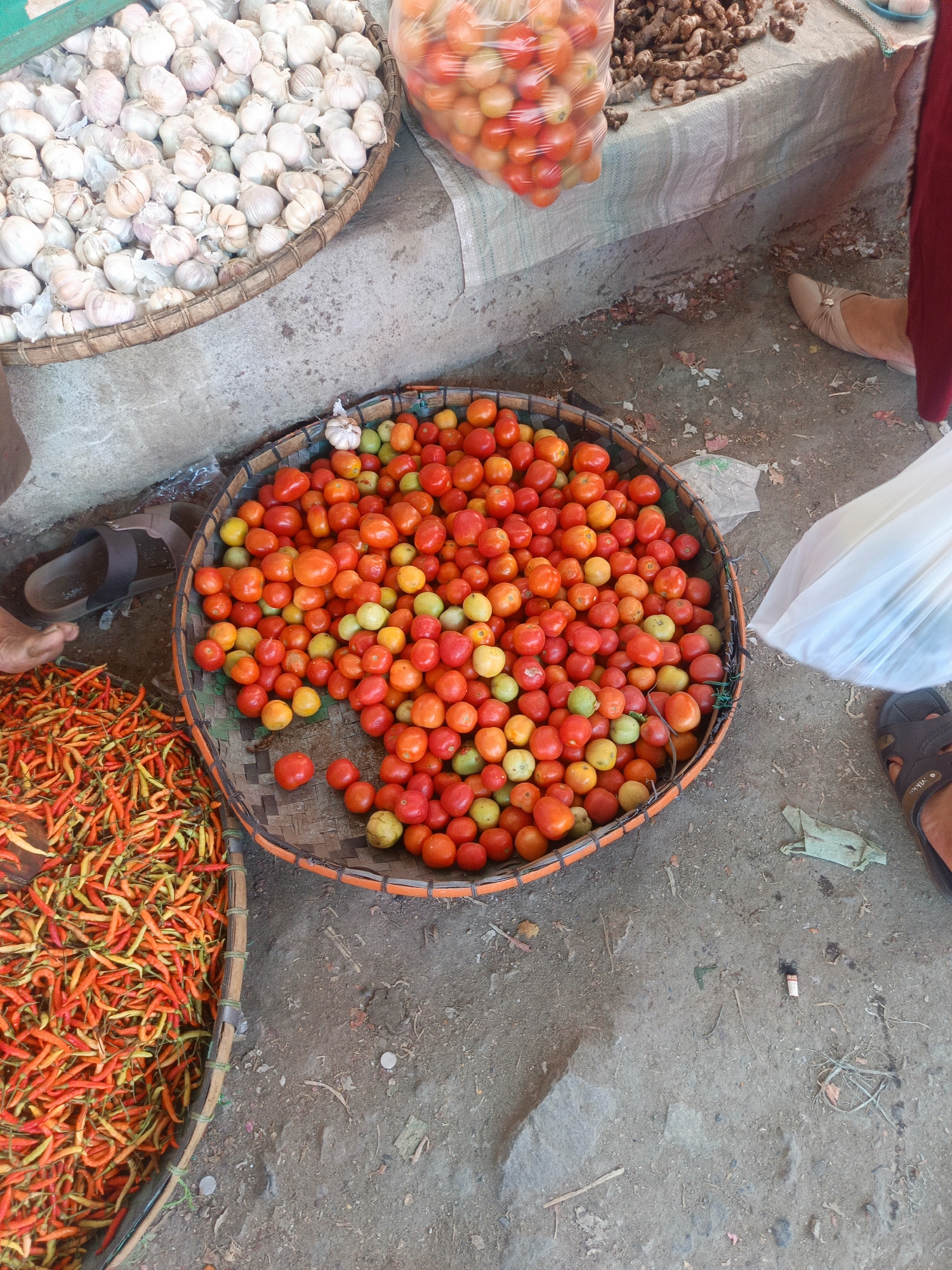 A top-down, medium shot captures an outdoor market scene in Gorontalo, Indonesia. In the foreground, a large, shallow basket overflows with vibrantly colored red and yellow chilies. To the right of the chilies, a round, woven basket is filled with a plentiful mound of ripe tomatoes, some appearing slightly green. Behind the tomatoes, a white plastic bag is held by an unseen person. To the left of the chilies, a partial view of a person's legs and feet, wearing a sandal, is visible. Further back and to the left, a woven basket holds a large quantity of white garlic bulbs. In the background, out of focus, a rough table displays more produce, including a plastic bag filled with tomatoes and what appear to be ginger roots. The ground is a mix of dirt and scattered debris.