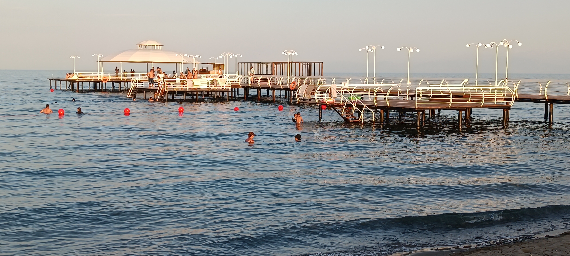 The image depicts a pier extending into a calm body of water, with several people swimming and standing on the pier. The pier features white railings and is supported by wooden posts. There are also lights and a covered seating area on the pier. Red buoys are visible in the water. The sky is clear and a light color, suggesting either early morning or late afternoon. This scene appears to be located at a beach or resort area.