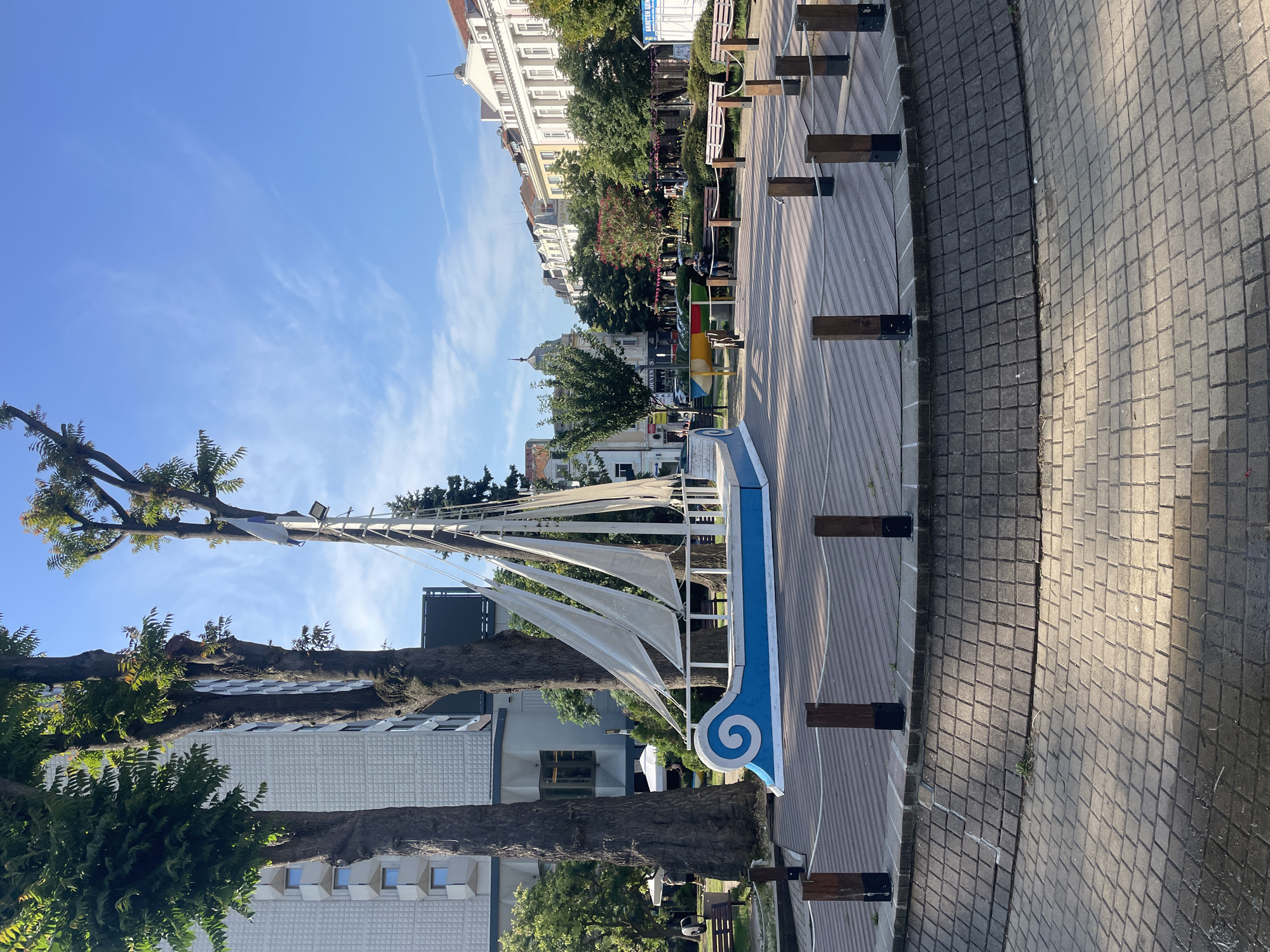 The image depicts a sunny day in a public square in Burgas, Bulgaria. The focal point is a decorative sailboat sculpture with white sails and a blue hull featuring a white spiral design. The sculpture is situated on a raised, circular wooden deck surrounded by a low wall made of grey bricks. The deck is further bordered by a paved area with a pattern of interlocking bricks, and wooden posts with ropes divide the space.
In the background, several large trees with green foliage rise prominently. Behind the trees, a modern, multi-story building with a textured white facade and square windows is visible. Further back, a row of older, European-style buildings with ornate architecture and mansard roofs lines a street. Some of these buildings appear to be shops or businesses, with signs visible.
Scattered throughout the square are several wooden park benches. A few people are present in the background, some sitting on benches, and others walking. One person is seated on a bench in the middle ground, looking towards the buildings. The sky is a clear blue with a few wispy white clouds. The overall environment suggests a pleasant, urban outdoor space.