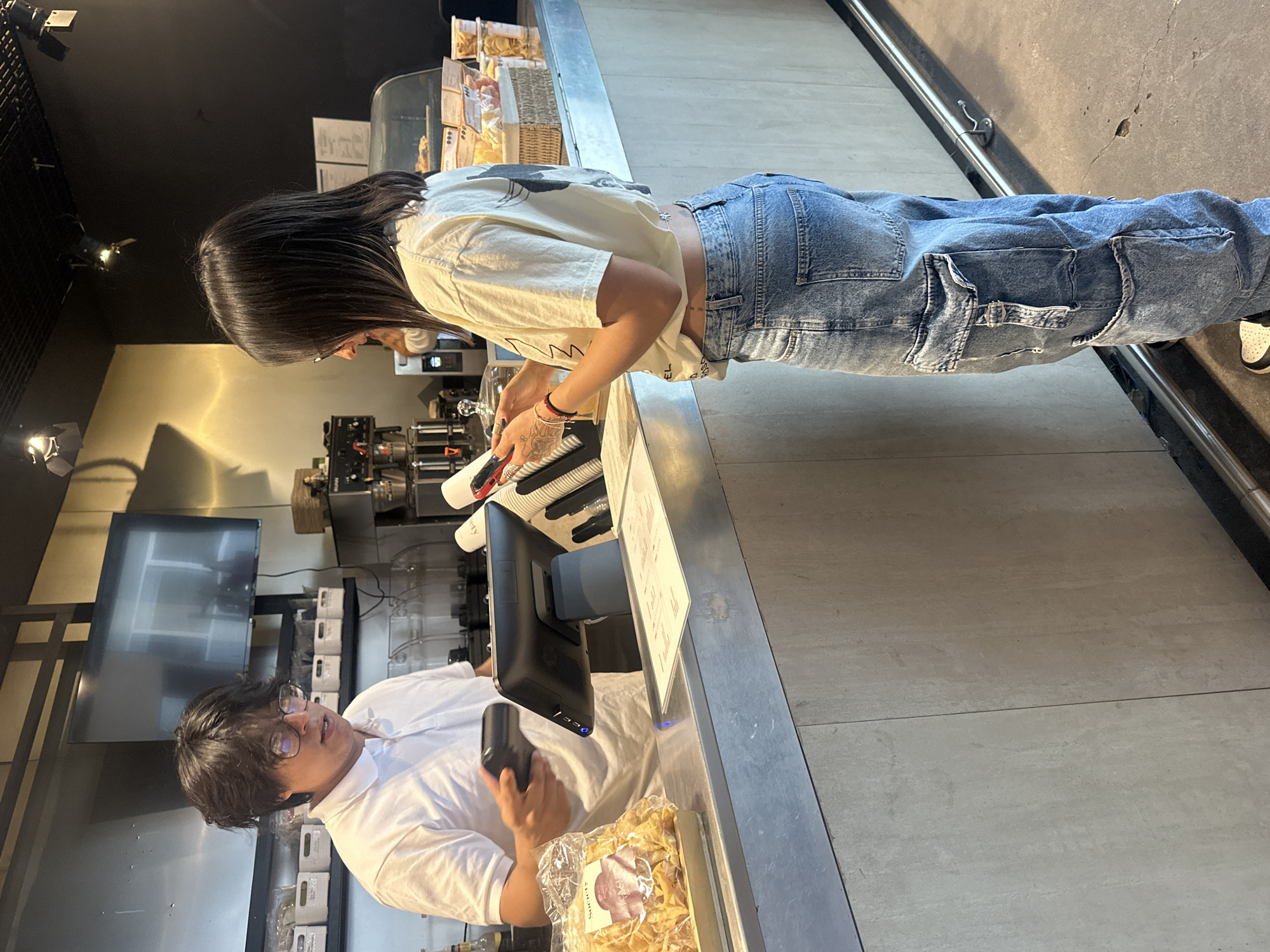 The image shows a cashier and a customer at a cafe or restaurant. The customer, a young woman with long dark hair, is wearing a light-colored t-shirt and blue jeans, standing at the counter and looking at something the cashier is showing her. The cashier, a young man with glasses and dark hair, is wearing a white shirt and holding a handheld device, seemingly processing a transaction. A cash register monitor and a menu are visible on the counter. In the background, shelves display various food items and supplies. The presence of a "Society" branded item suggests this could be a location of Society Cafe in Mexico City, Mexico, given their presence there. The overall environment suggests an indoor setting with modern decor and food retail activity.