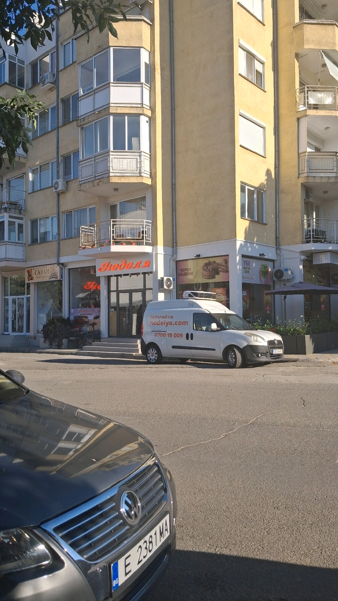 In the city of Gotse Delchev, Bulgaria, a street-level view captures the facade of a building housing multiple establishments. On the left, a shop labeled "САНКИ" (SANKI) with text below it suggests it is a shop selling something related to winter or children's items. Next to it, a prominent establishment displays the name "Неделя" (Nedelya) in red illuminated letters above its entrance, indicating a business that is likely a bakery or cafe, as suggested by the signage visible on the van.
A white delivery van is parked in front of "Неделя," bearing the website "NEDELYA.COM" and a phone number "0700 19 009" in red text, indicating it's a vehicle for the establishment. To the right of "Неделя," another shop displays images of cakes and pastries, with text that includes "ЧЕРНО КАДИФЕ" (CHORNO KADIFE - Black Velvet) and "УБЕ МАТЧИ" (UBE MATCHI). The environment appears to be a sunny day, with shadows cast on the pavement and building. A gray Volkswagen car is partially visible in the foreground, with its license plate reading "E 2381 MA." The building itself is a light yellow color, with multiple floors and balconies. The overall scene depicts a commercial street in a Bulgarian city.