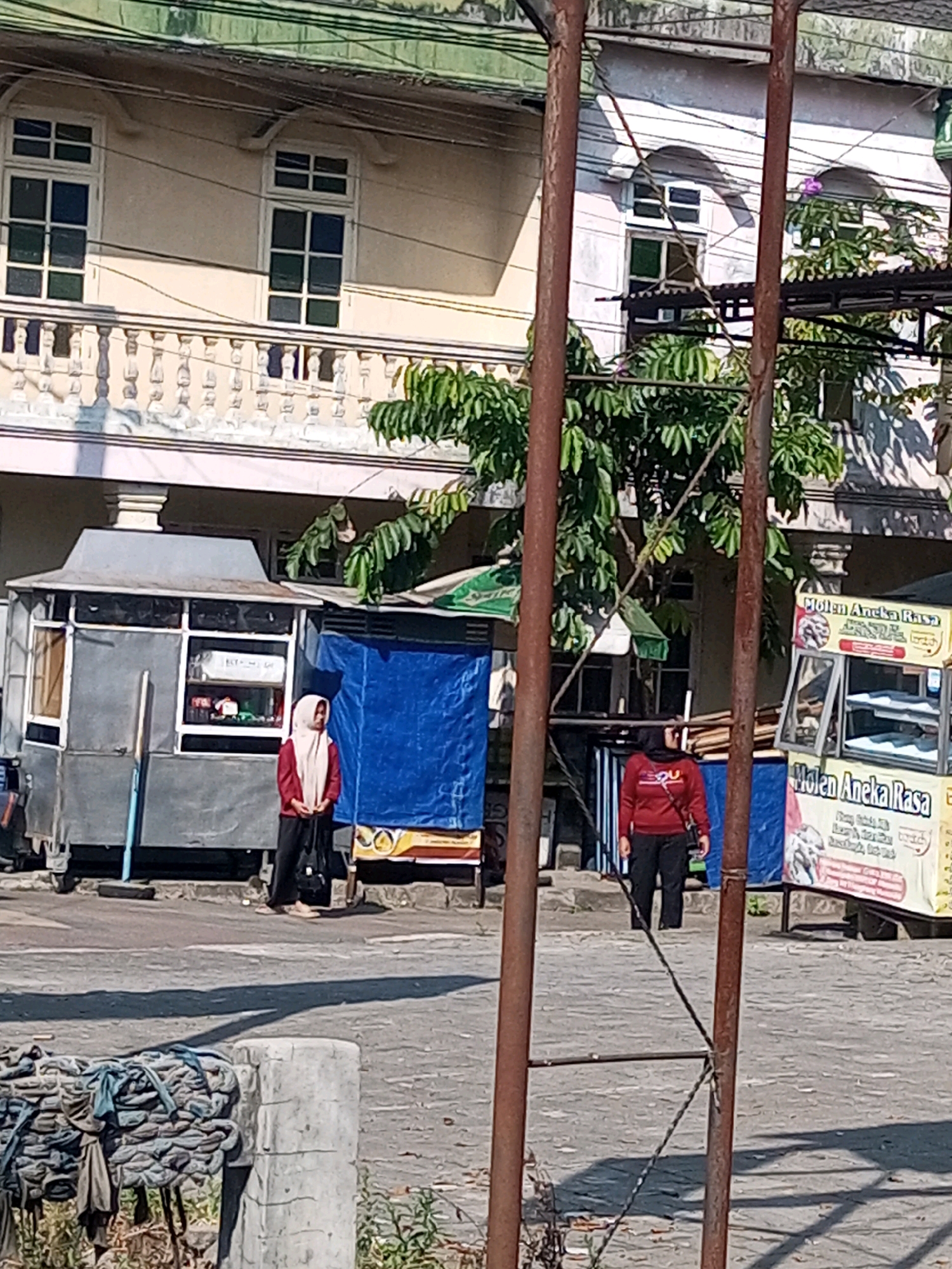 The image captures a street scene in Wonosobo, Indonesia, on a bright, sunny day. In the foreground, a thick, rusty metal pole stands prominently, with a thin wire attached. To the left, a substantial pile of tightly bound ropes rests on a concrete cylindrical base. Behind this, a gray street cart with a metallic roof and transparent windows is visible. A woman wearing a light pink hijab and a dark red jacket, with black pants, stands to the left of the cart, facing right. Further to the right, another cart, predominantly yellow with blue and red accents, displays signage that reads "Molen Aneka Rasa" and features images of food items. A woman in a red long-sleeved shirt and dark pants, also wearing a headscarf, stands to the right of this cart, facing left. In the background, a two-story building with a light yellow facade features a balcony with ornate white railings on the upper level. The upper level also has multiple windows with white frames and green-tinted panes. Trees with dense green foliage are interspersed between the carts and the building. The ground is paved, and shadows are cast across it, indicating the strong sunlight.