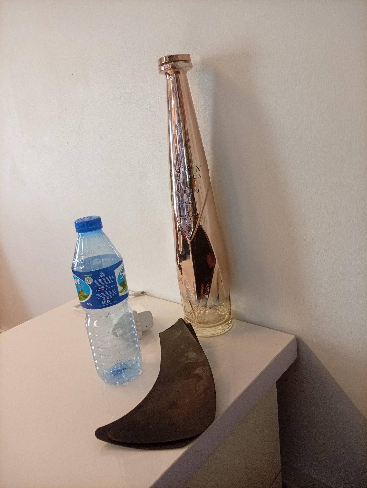 A water bottle, a bronze colored bottle, and a pair of dark brown shoe inserts are sitting on a white surface. The bottles are positioned against a white wall. The scene is indoors. There is no discernible information to associate this scene with Jos, Nigeria.