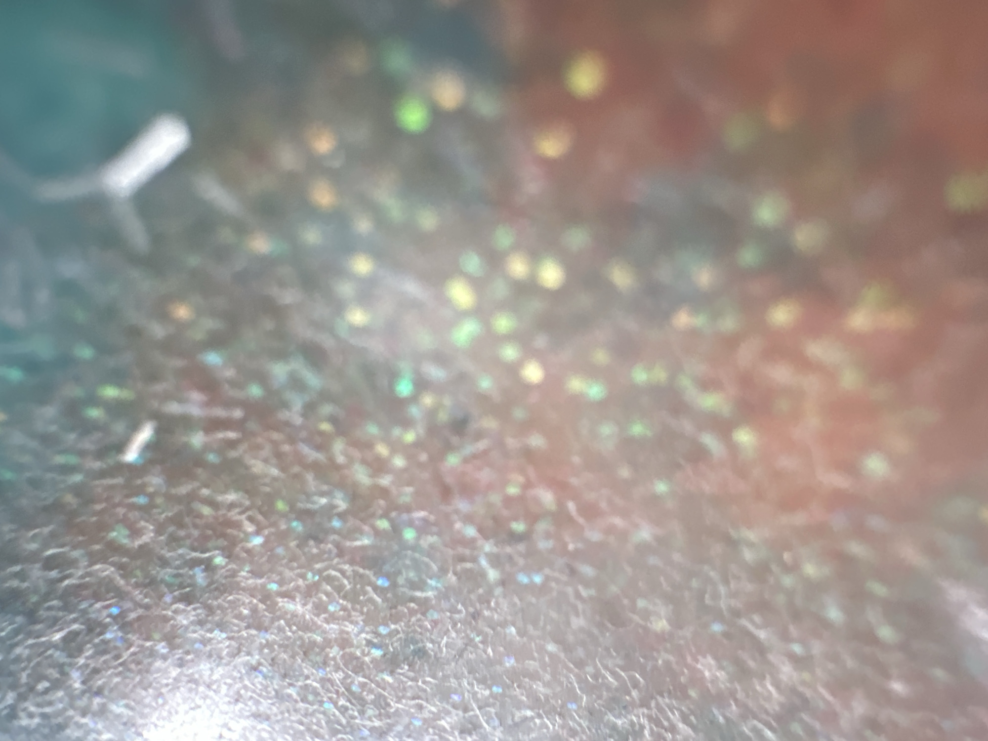 This is a close-up, out-of-focus shot of a textured, holographic surface. The surface appears to be a material that reflects light, creating a shimmering effect with various colors, including blues, greens, yellows, and pinks. The texture is somewhat rough and uneven, with small, raised areas or particles. In the upper left corner, a blurry white object, possibly a cylindrical piece of plastic or metal, is visible with two faint lines extending from it, suggesting it might be an attachment or a tool. The lighting creates specular highlights and bokeh effects, making it difficult to discern a specific scene or setting. The image does not contain any discernible text, people, or clear indicators of time of day, weather, or specific location cues, beyond the provided metadata indicating Belgrade, Serbia. The overall impression is abstract and decorative, focusing on the interplay of light and texture.