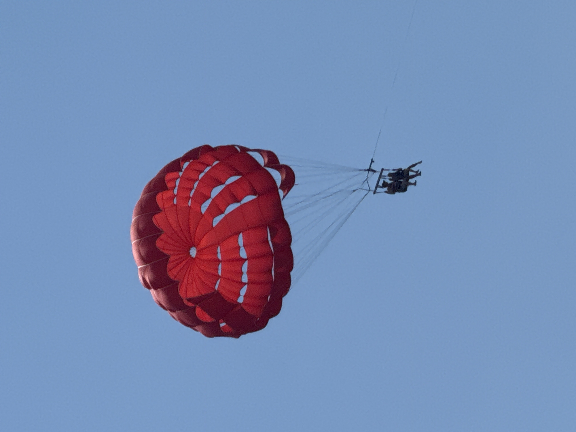A low-angle shot depicts two people parasailing under a red parachute against a clear blue sky. The figures are small and silhouetted against the sky, positioned to the right of the frame. The parachute is large, inflated, and features a radial pattern. The background is a uniform blue. The location is identified as Budva, Montenegro, based on the presence of parasailing activities.