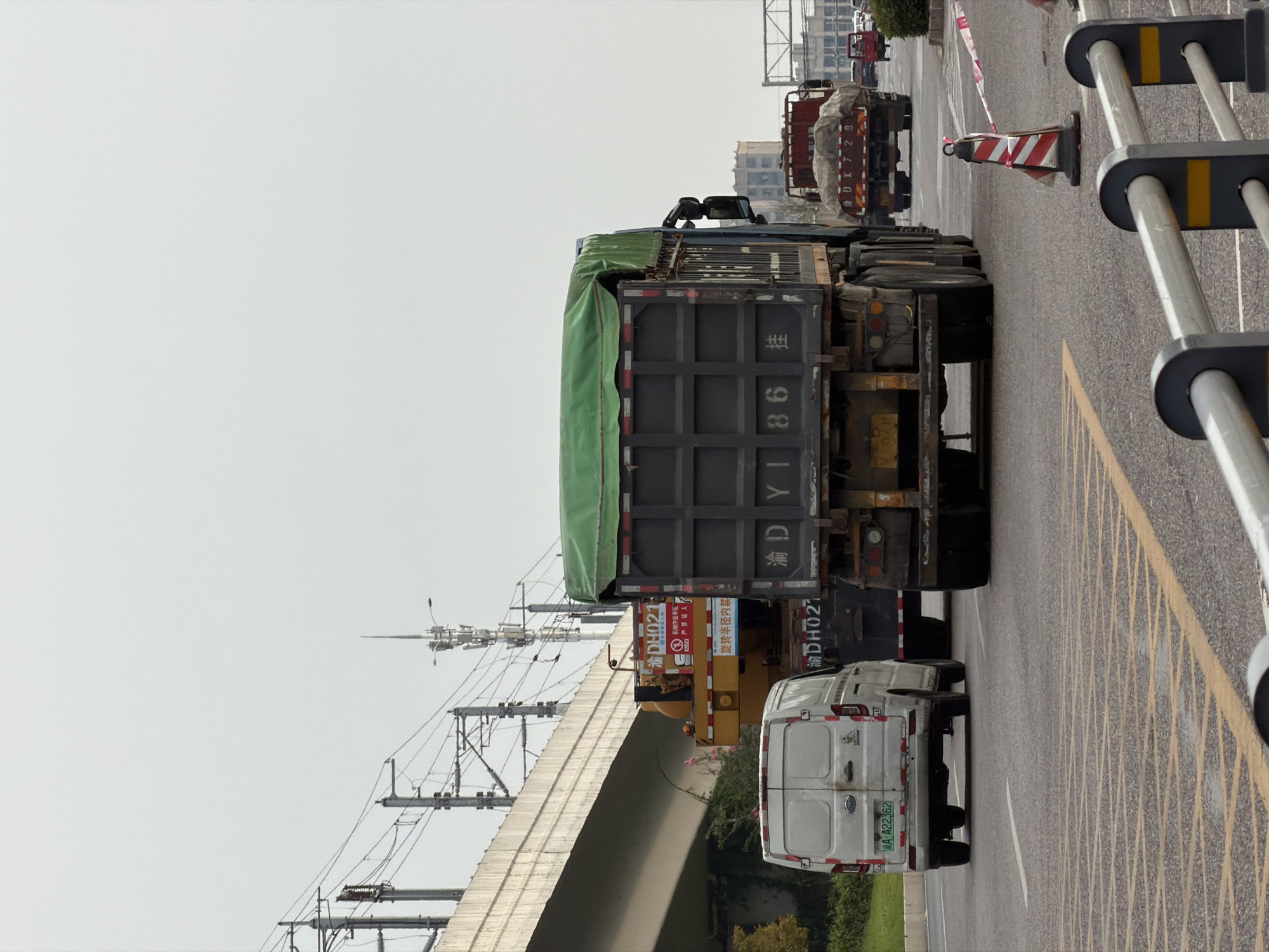 The image shows a street scene in Jiangjin, China, with several vehicles present. In the foreground, on the right side of the road, there are construction barriers with red and white striped cones and tape, indicating a potential work zone or traffic diversion. In the center of the image, a large dark gray dump truck with a green tarp covering its load is prominently featured. The truck has "Y DY 1 86 挂" written on its back. To the left of this dump truck, a white van is visible, with a license plate reading "A A22362". Behind the white van, a yellow construction vehicle with some signage on its side is partially obscured. Further down the road, another red dump truck with a covered load is visible, and beyond that, a smaller red vehicle is seen. Overhead, power lines and infrastructure suggest an urban or semi-urban setting. The sky is overcast. The road surface has visible yellow lane markings and some gravelly texture.