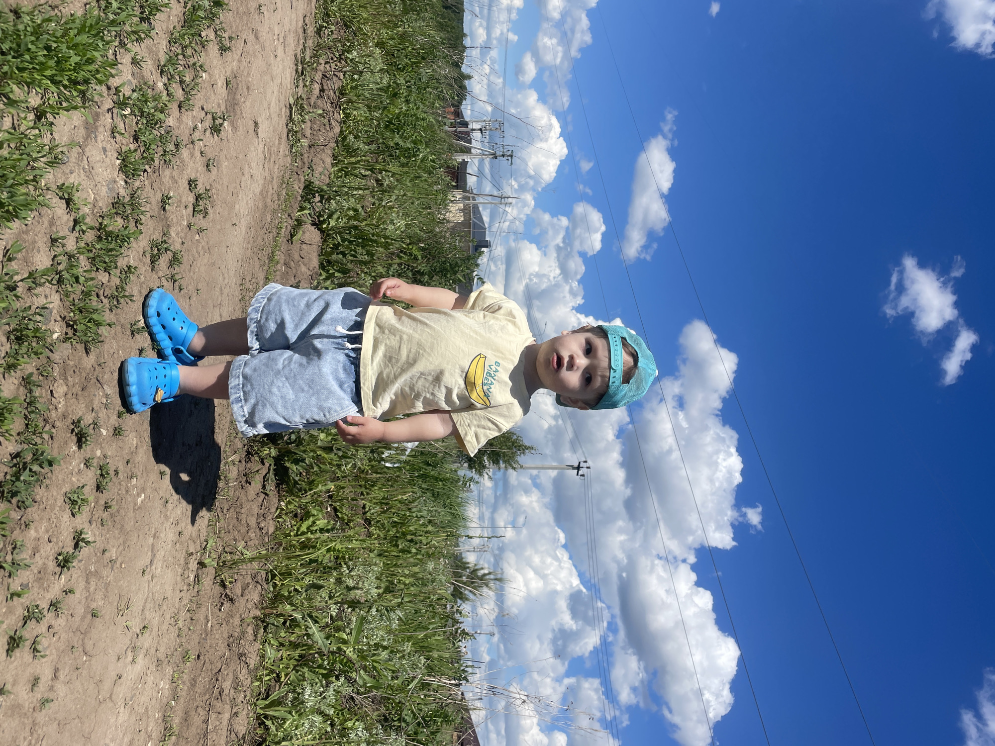 A young child stands on a dirt path lined with green vegetation, beneath a vibrant blue sky dotted with fluffy white clouds. The child is wearing a yellow t-shirt with a banana graphic, light blue shorts, bright blue Crocs shoes, and a blue baseball cap. Power lines are visible extending across the sky behind the child. The location appears to be Эстачи, Russia based on visual cues. The child is looking towards the camera.