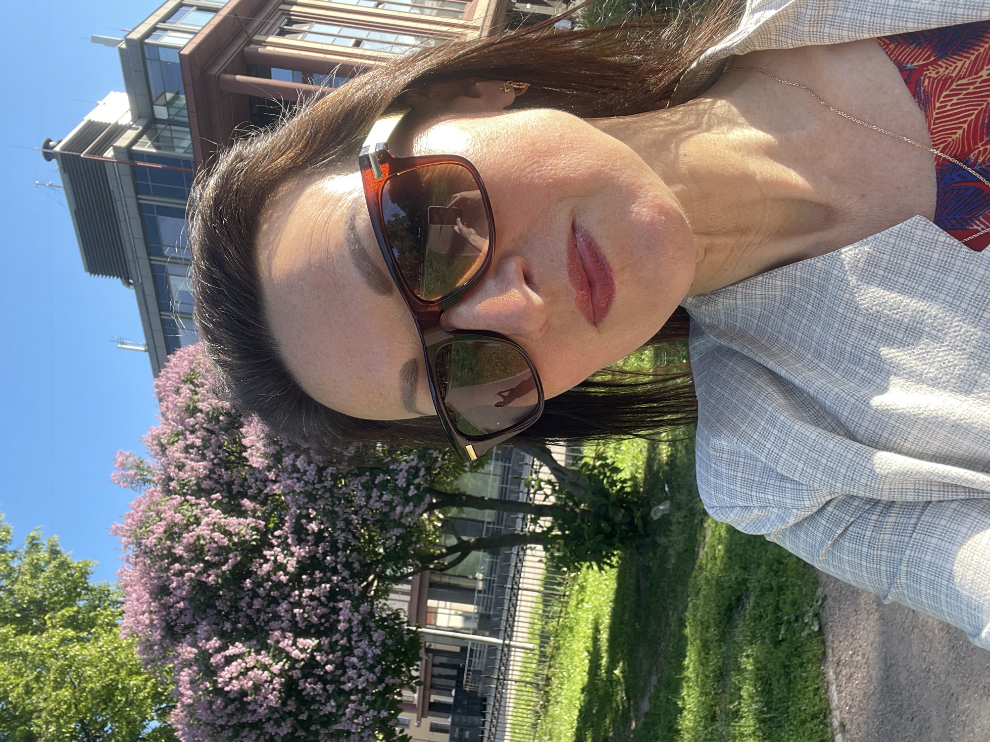 A woman wearing sunglasses and a light-colored blazer takes a selfie outdoors. She has dark brown hair and is wearing lipstick. In the background, there is a flowering bush with pink blossoms, a grassy area, a building with a light brown facade and large windows, and a bright blue sky. The scene appears to be in a city park or urban green space.