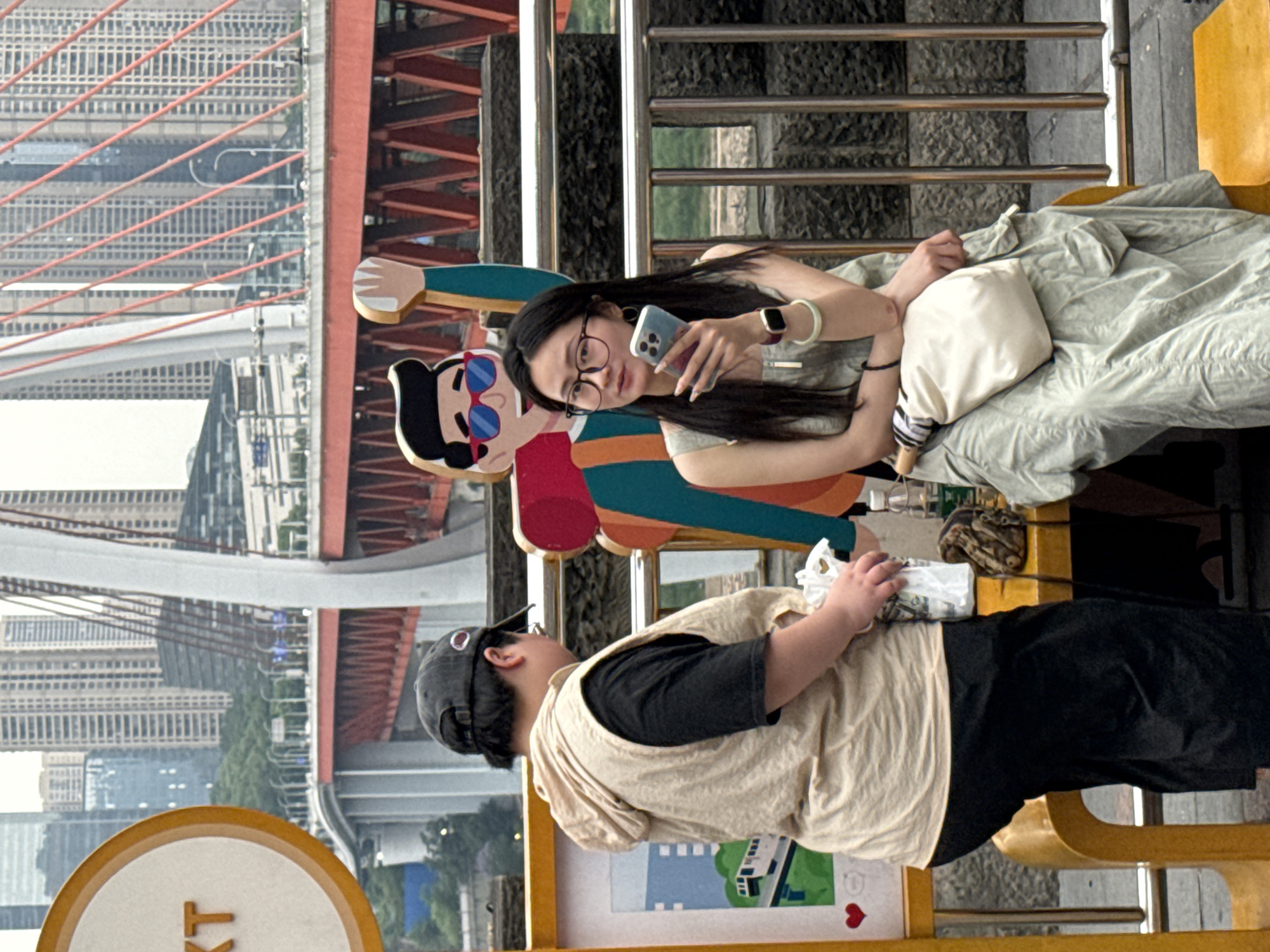 In Yuzhong District, China, a horizontally composed image captures two individuals in an outdoor setting. A woman with long dark hair and glasses is holding a smartphone, seemingly taking a photo. Adjacent to her, a cardboard cutout of a male figure adds a playful element to the scene. In the foreground, another person is present, their features partially obscured. The backdrop showcases urban architecture, including a bridge and buildings, indicating a city location. Additional elements include a blanket, a purse, a sign with a heart symbol, and metallic railings, adding depth and context to the composition.